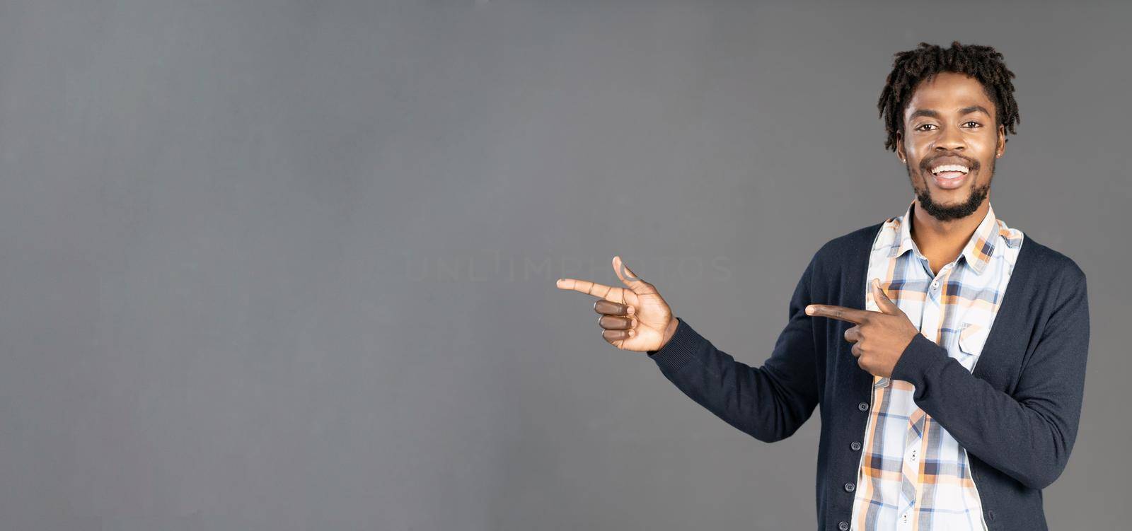 Pointing or directing young african american man in dark navy jumper with both hands turn sideways looking at camera and smile, isolated on grey background. Elegant male model in studio. copy space