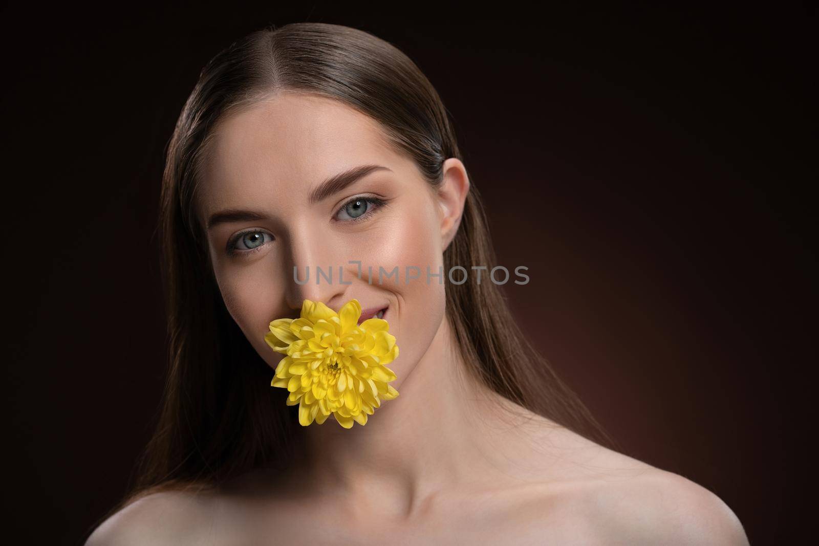 Smiling beauty model with bare shoulders holding a flower in her mouth with a beauty skin and long hair looking at camera isolated on black background. Woman nature cosmetic spa concept.