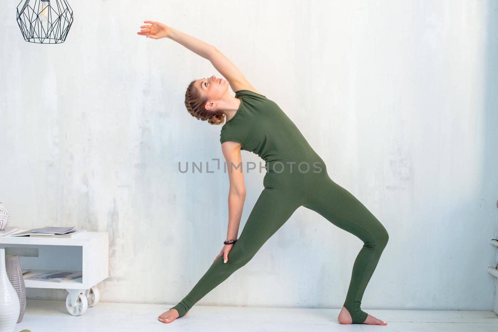 A slender beautiful woman in a green sports jumpsuit, near a light wall, performs a yoga exercise