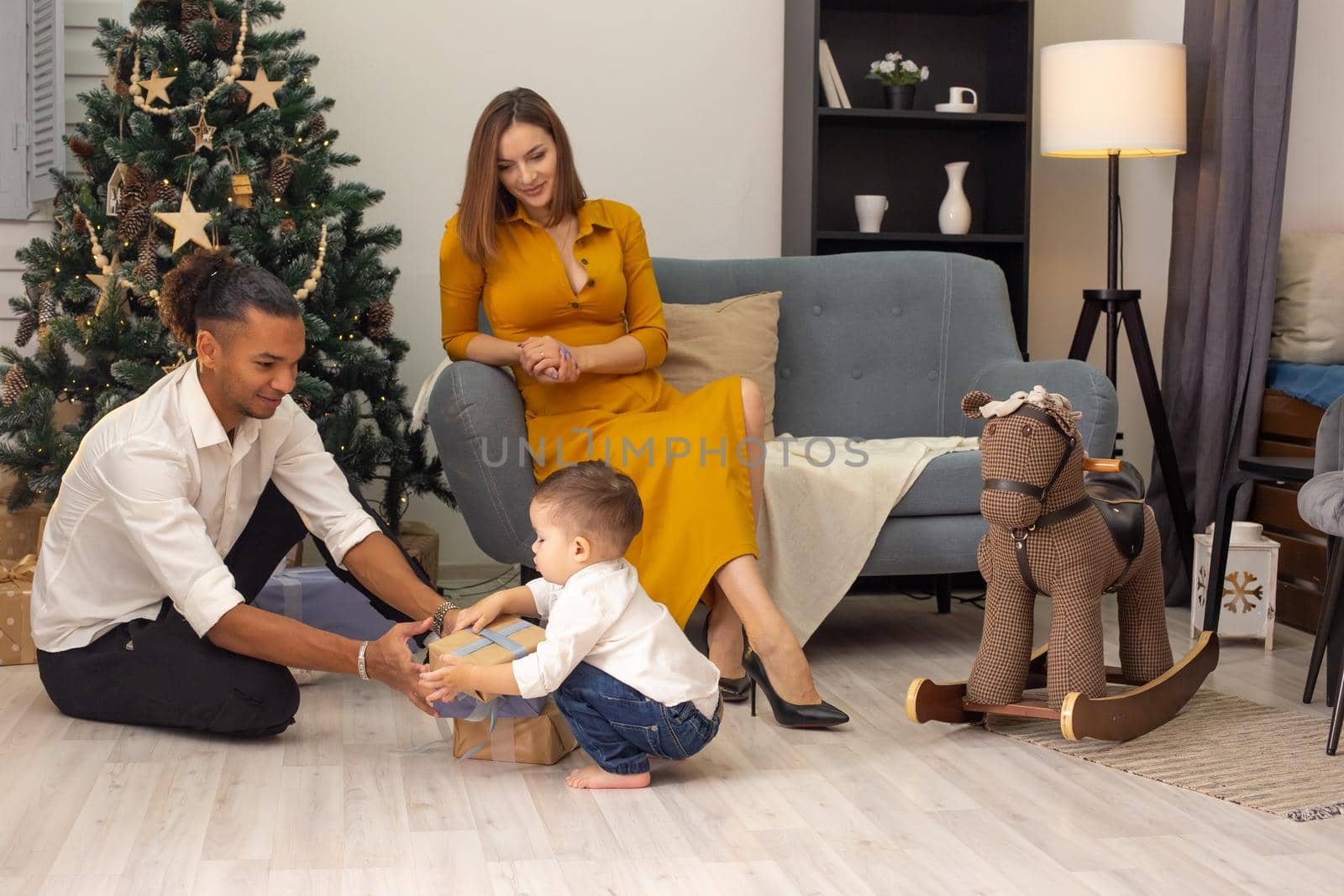 A beautiful slender girl in a mustard dress sits on a gray sofa, a young man and a toddler child cheerfully sort out boxes with gifts, on the floor, near a Christmas tree