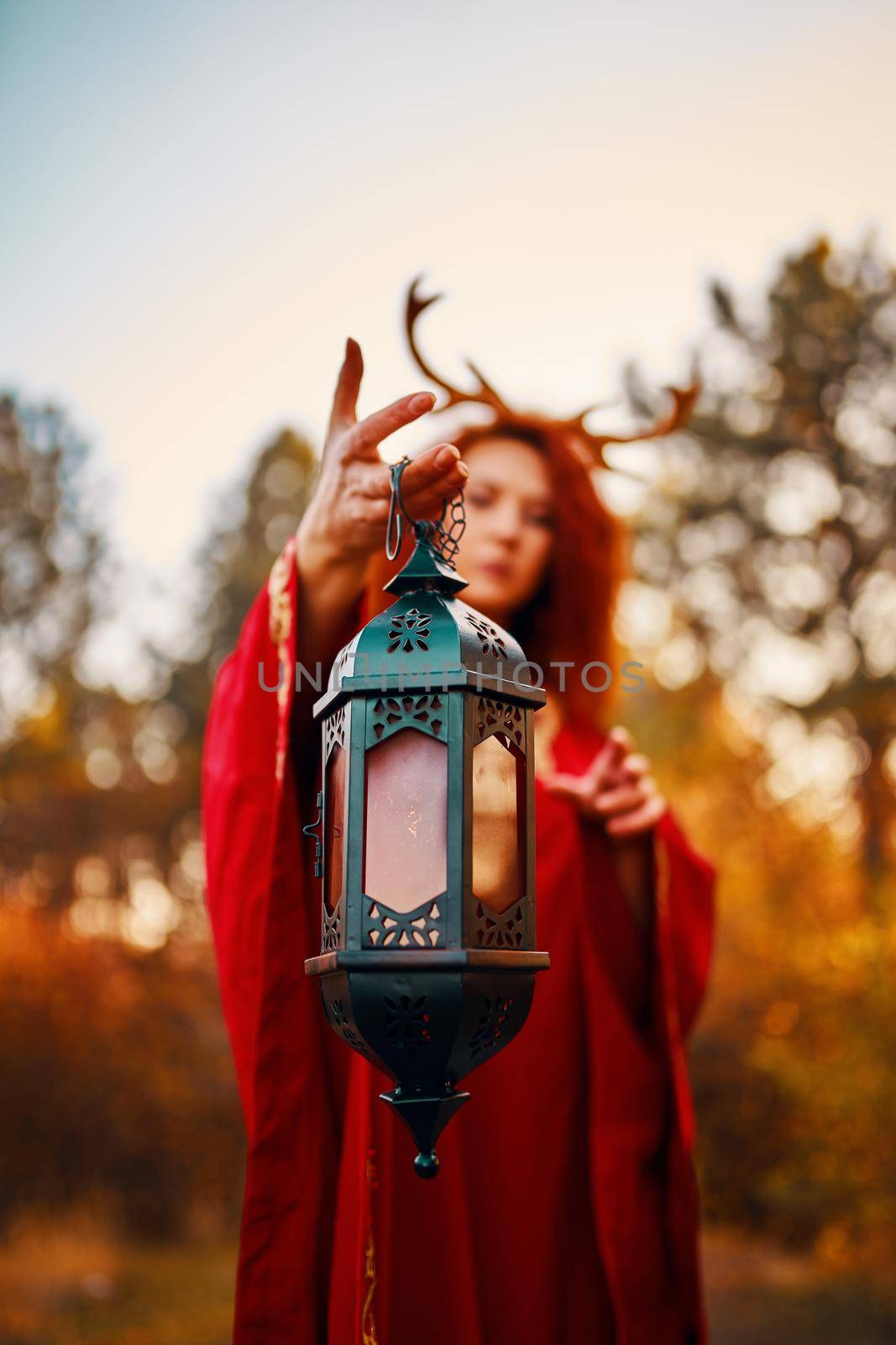 Woman in long red dress with deer horns in autumn forest. The demon of fairy tales in a red cloak with a lantern in his hands walks through the autumn forest and looks for magic herbs.