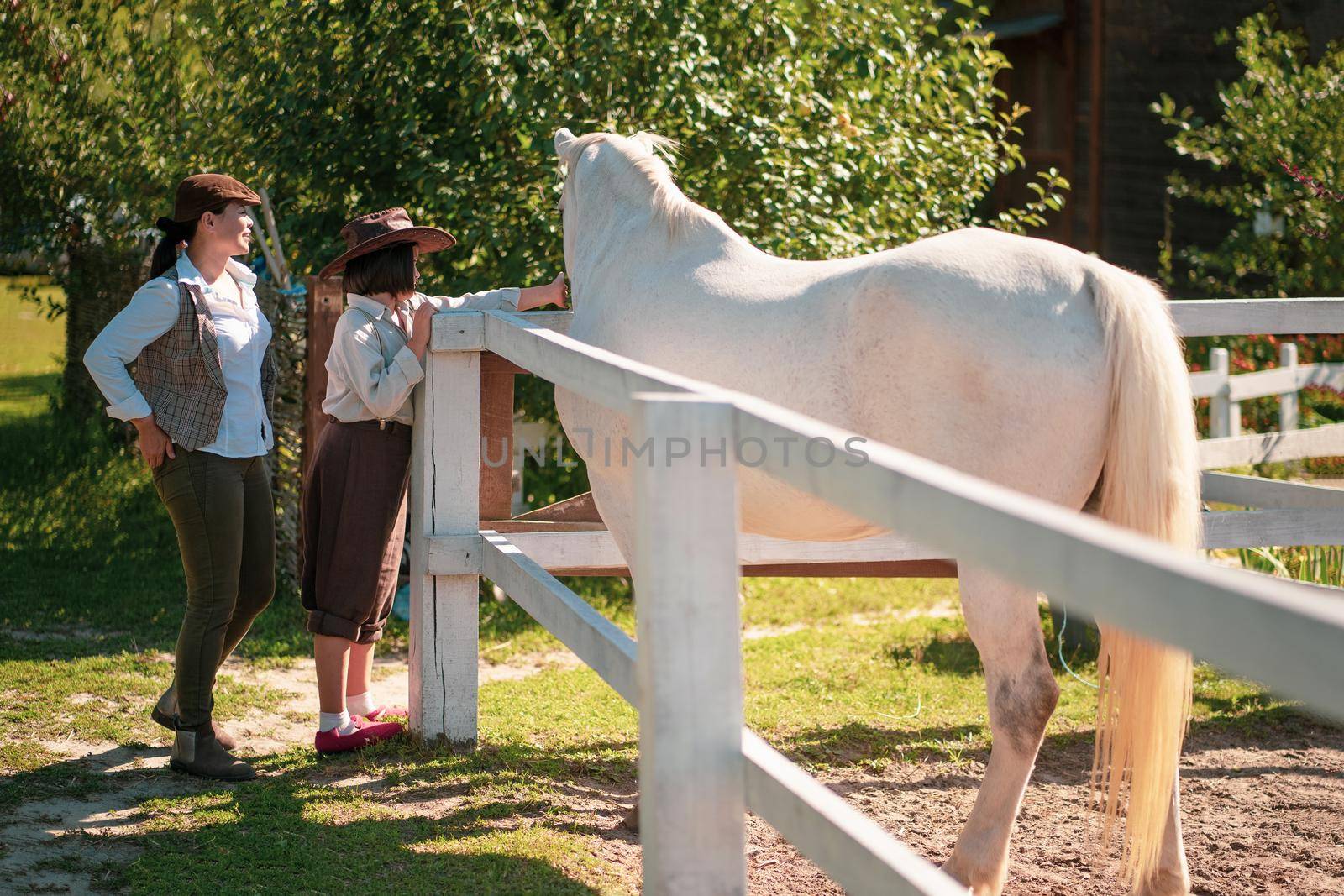 Happy farm living. Mother and daughter in vintage clothes stand near the paddock with a beautiful white horse. The girl is stroking a horse. Family at the ranch.