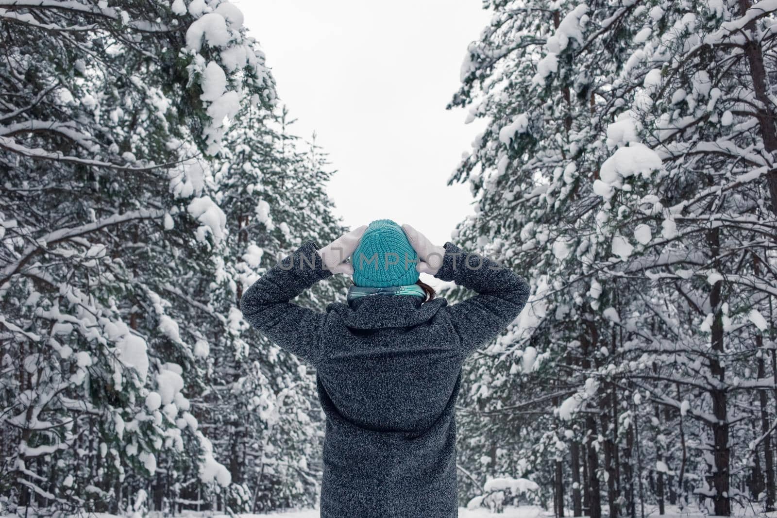 A girl in a gray coat and a light blue knitted hat stands with her hands to her head in a beautiful winter snowy forest on a cloudy snowy day. Back view