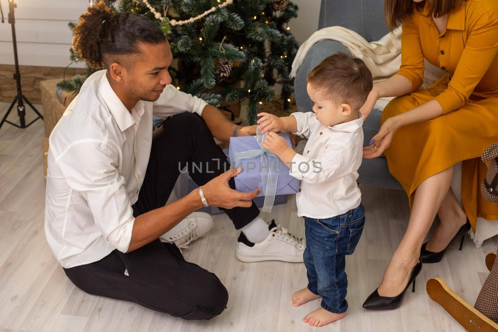 In the living room, near the Christmas tree, a stylish father sits on the floor, helping his little son unpack a box with a gift. Nearby sits a slender beautiful mother in a dress