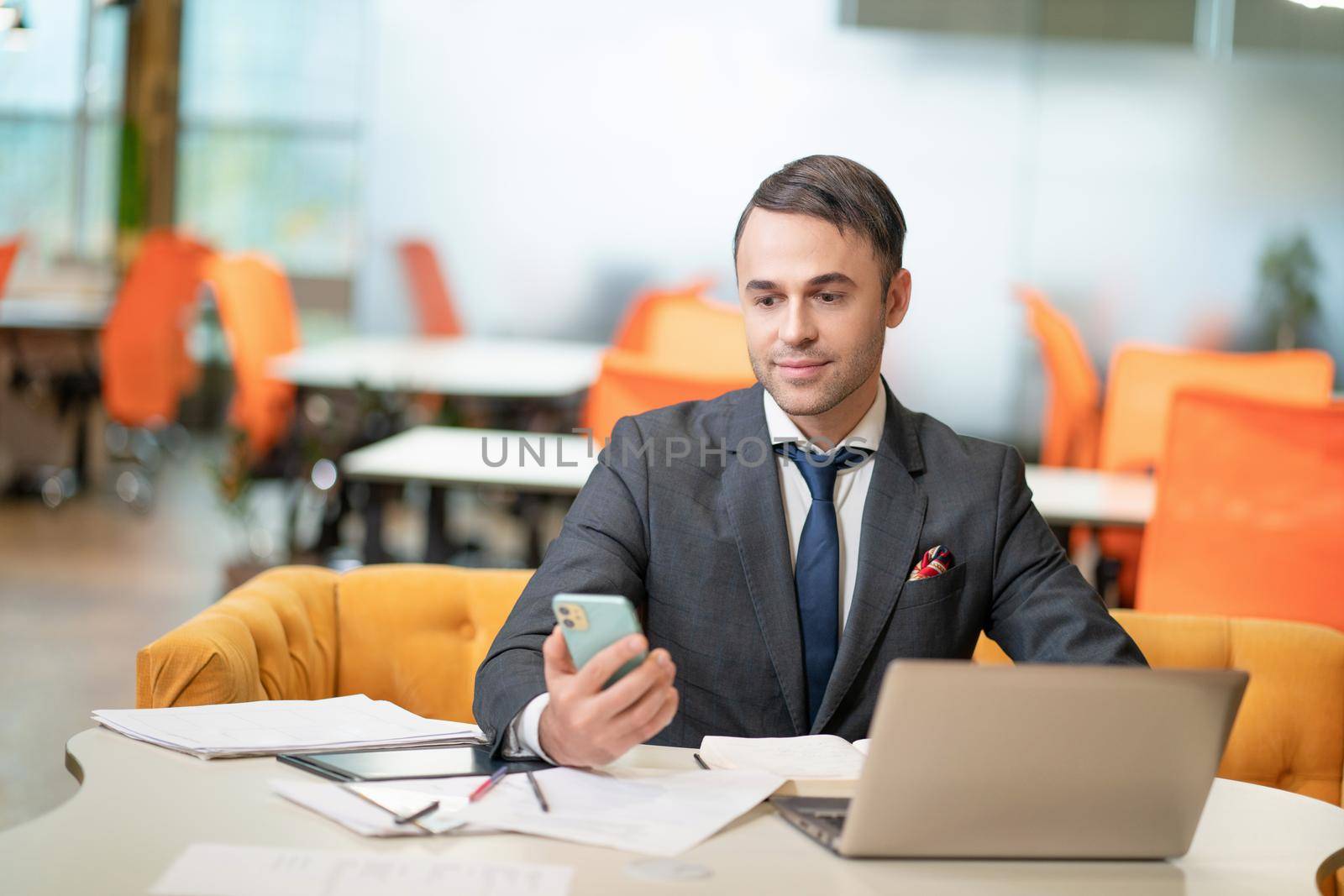 Young businessman or manager looking on phone while working on laptop sitting at the modern office with toxic orange chairs on background.