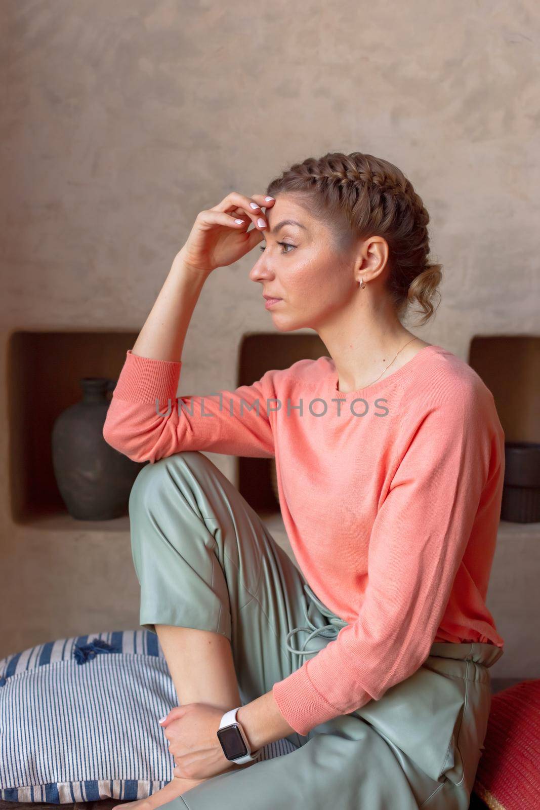 pensive beautiful woman in coral sweatshirt and gray pants is sitting in the room. Vertical