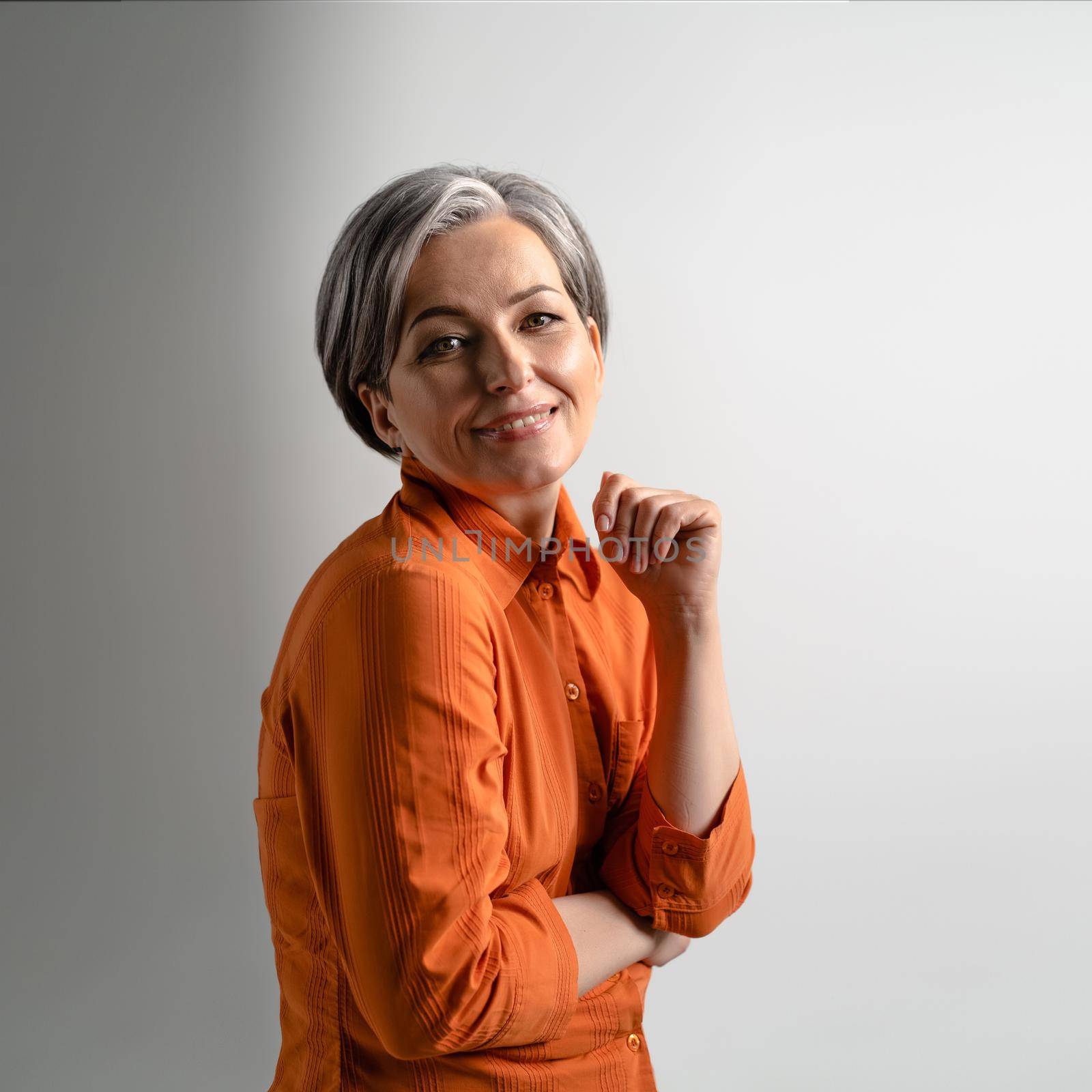Portrait of a mature grey haired woman wearing orange shirt happily smiling on camera. Pretty mid aged grey haired woman in orange shirt isolated on grey background by LipikStockMedia