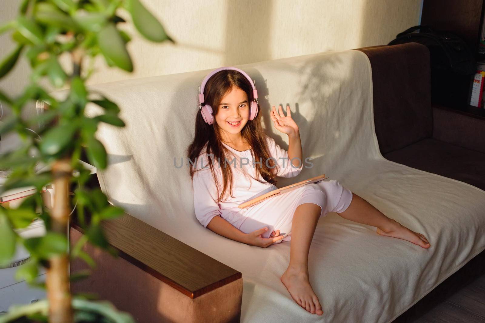 A friendly little beautiful girl in pink home clothes and pink headphones sits on the couch, holds a digital tablet in her hands, smiles, looks into the camera