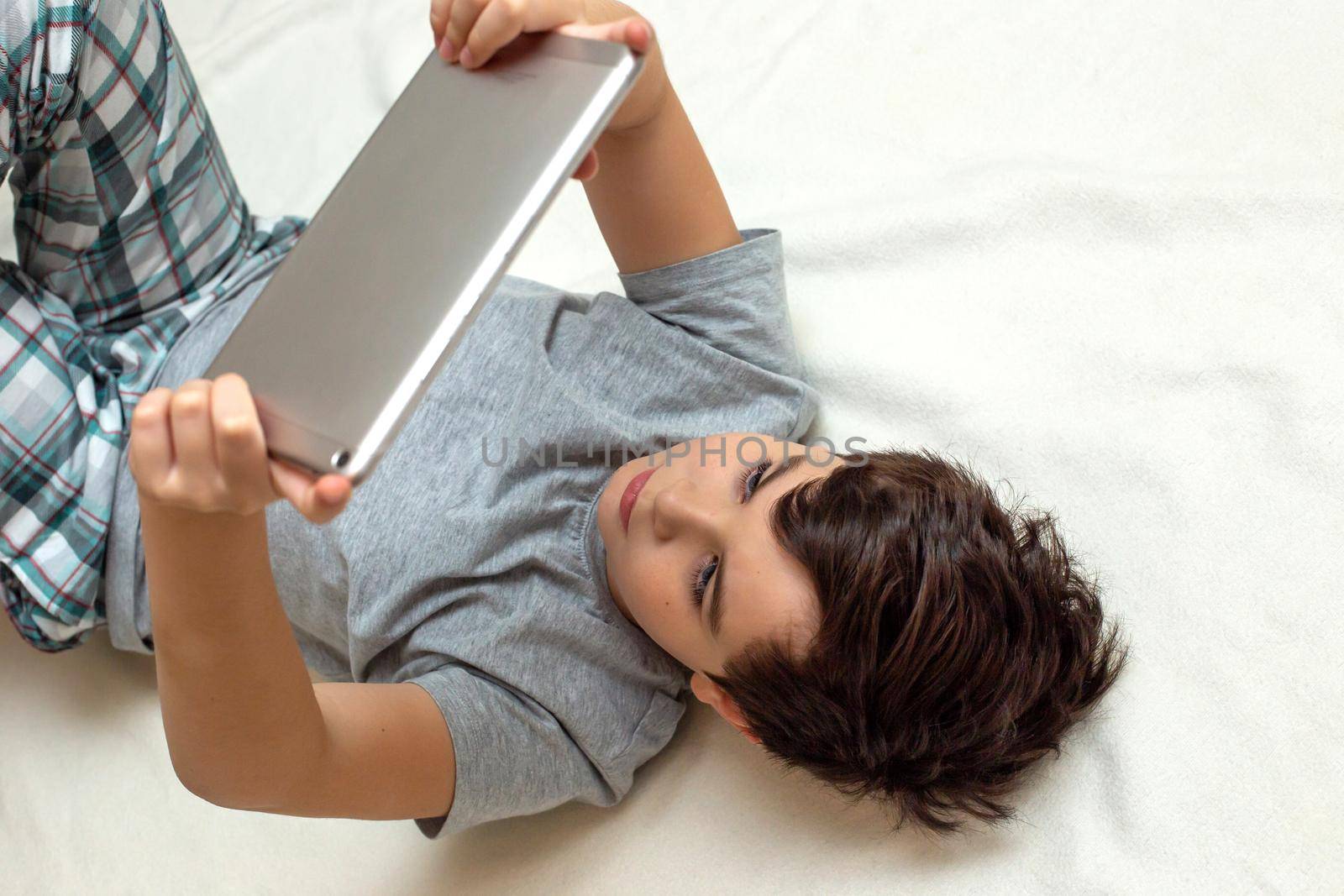 A brunette boy in gray pajamas , lies on his back on a white blanket and plays on a digital tablet. Copy space