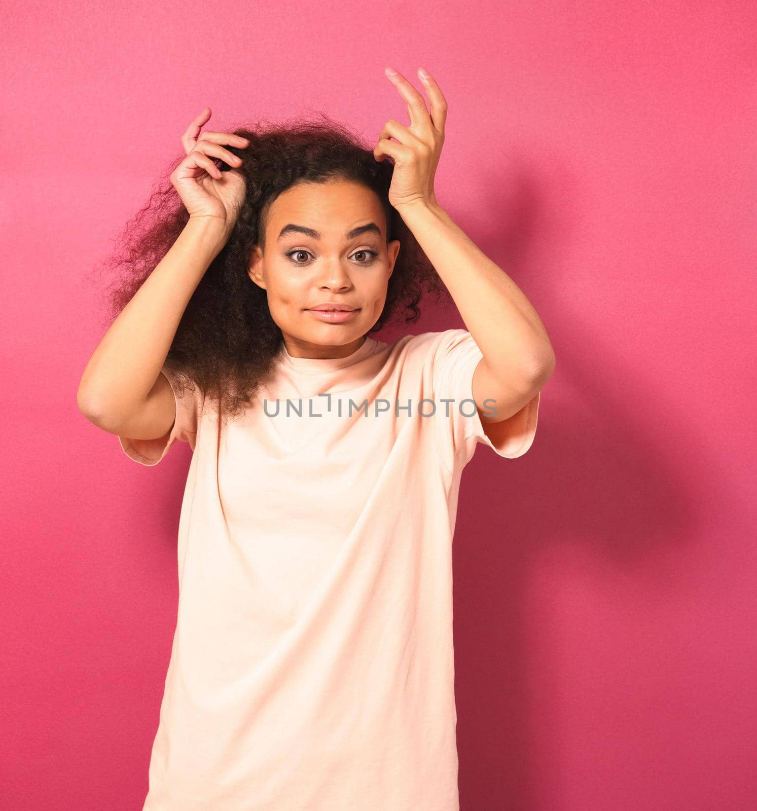 Beautiful young African American woman with Afro hair dancing looking positively at camera wearing peachy t-shirt isolated on pink background. Beauty concept.