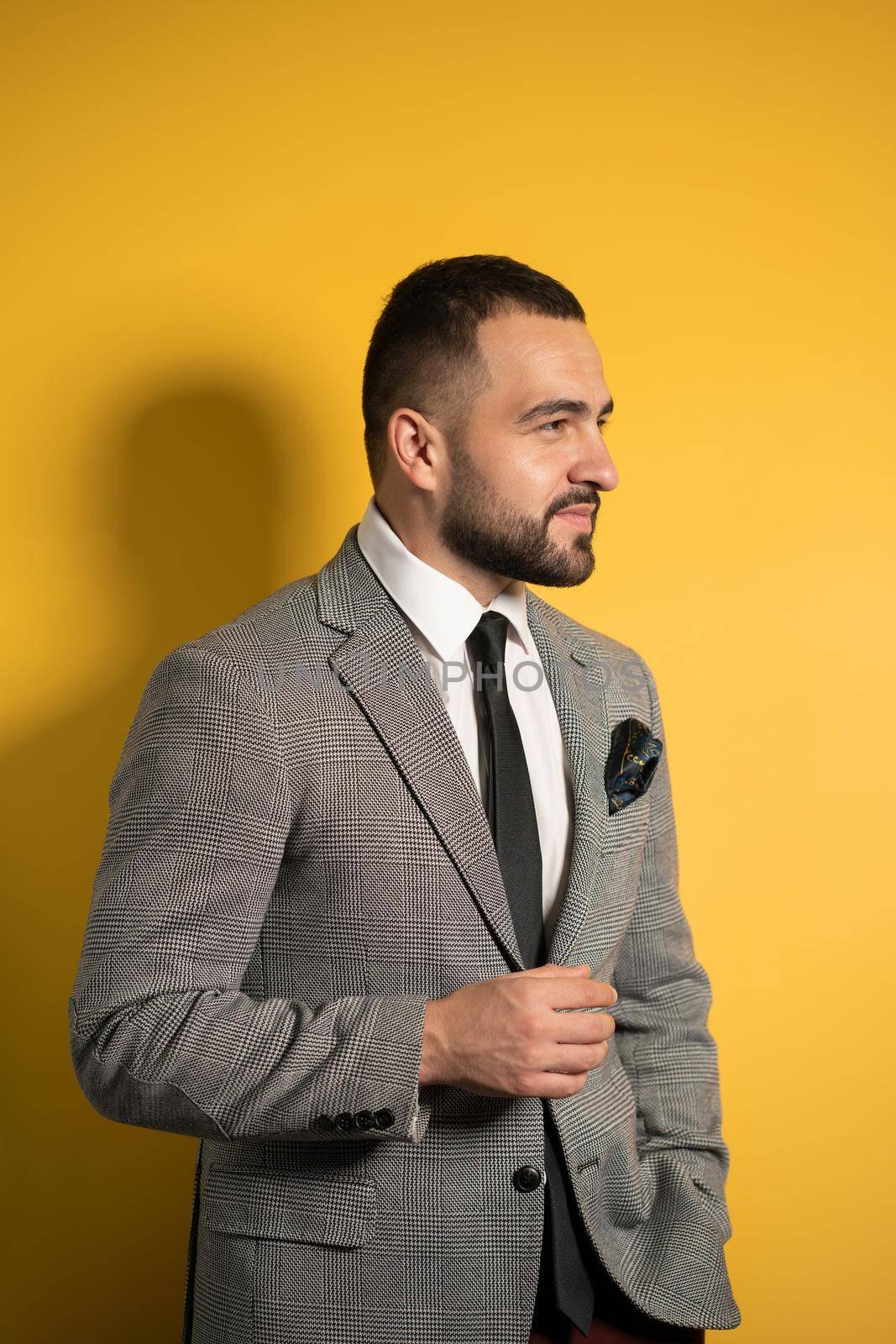 Handsome smiling young man wearing grey suit with one hand lifted standing half sideways isolated on yellow background.