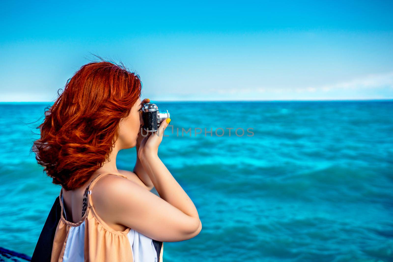 Red haired woman in summer dress travel photographer, taking pictures of sea. Rare photographic camera for vacation snapshots.