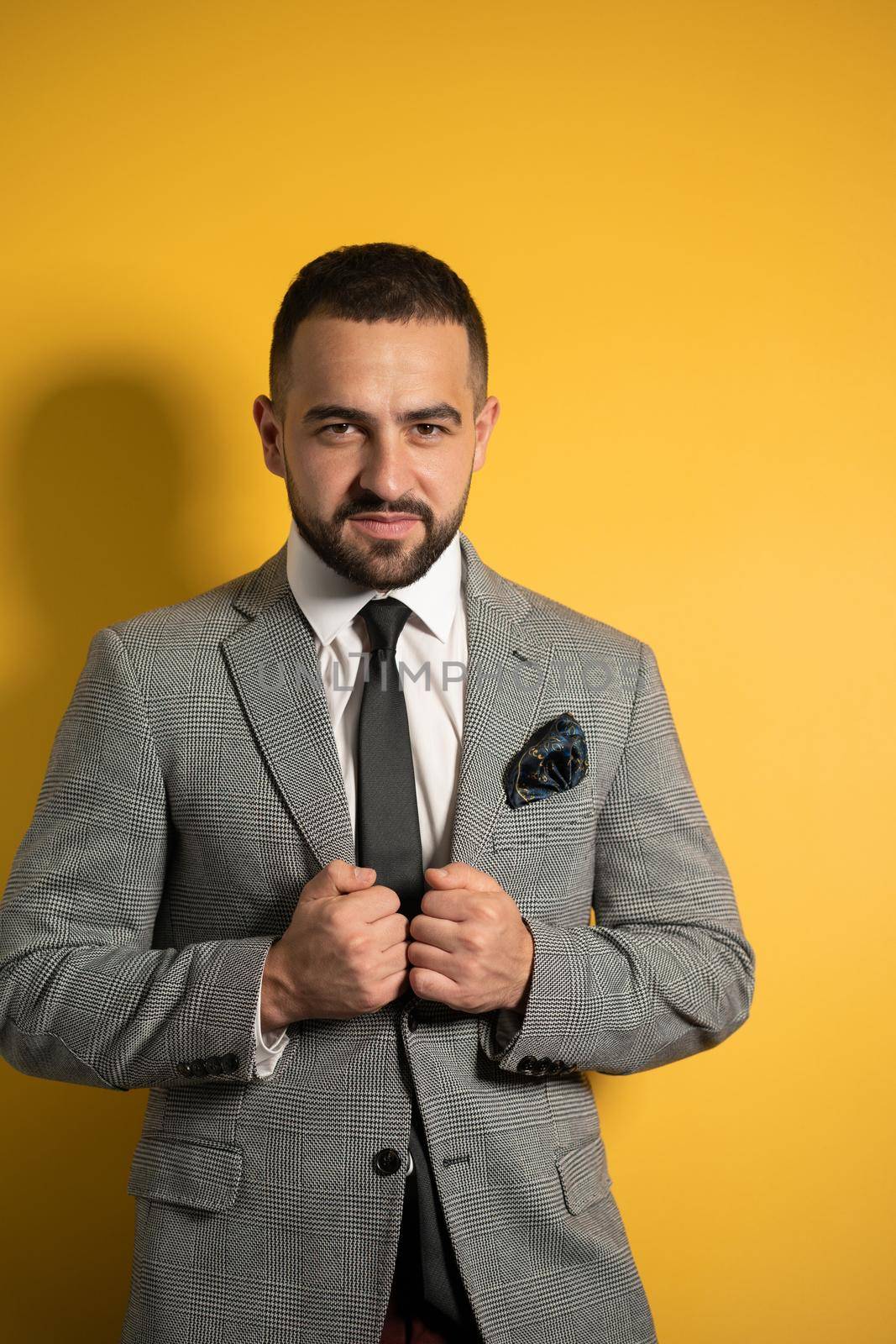 Holding jacket handsome slightly smiling young businessman wearing grey suit with one hand lifted standing half sideways isolated on yellow background.