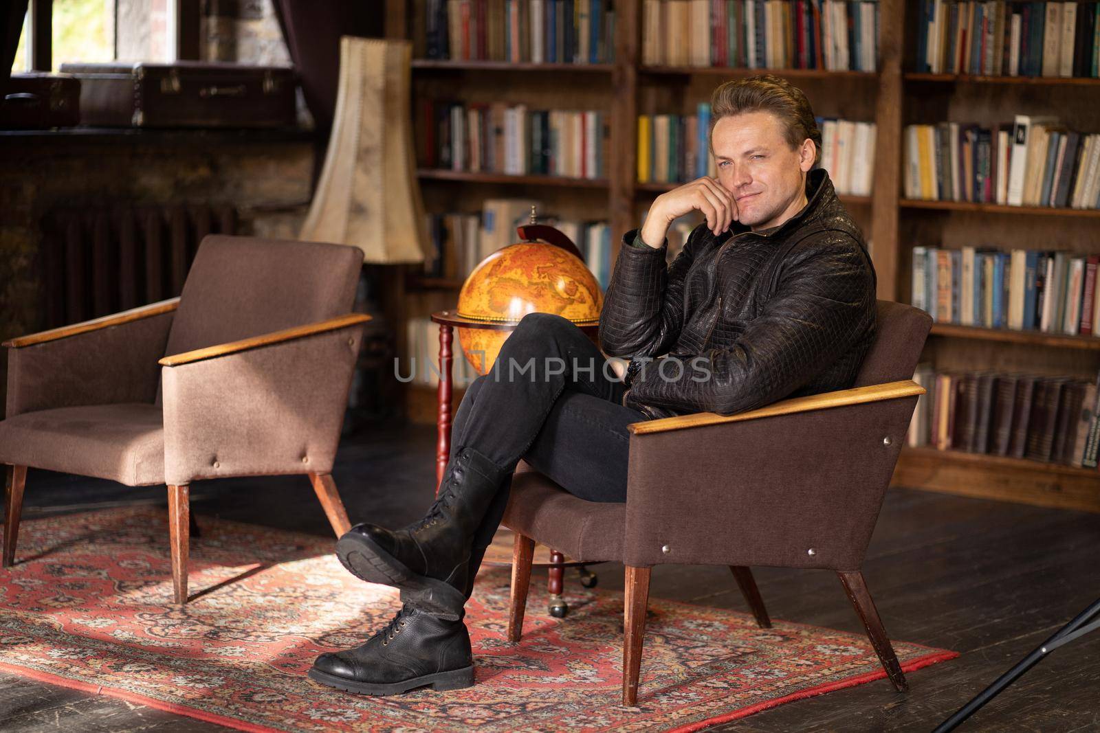 pensive man in a leather jacket and ankle boots sits cross-legged in an old armchair in a vintage room. Caucasian man sits in an armchair in the middle of an antique living room against the backdrop of shelves with books. Rustic rug on the floor. Close-up . High quality photo