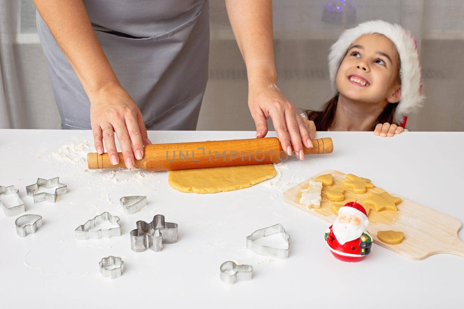 woman's hands in a gray apron, roll out the dough for Christmas cookies with a rolling pin on a white table. Funny little girl in a red cap peeks out from under the table