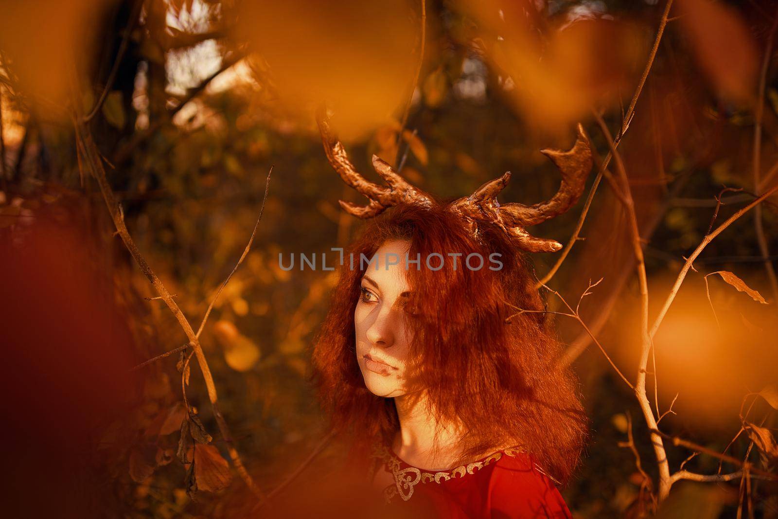 Woman in long red dress with deer horns in autumn forest. The demon of fairy tales in a red cloak with a lantern in his hands walks through the autumn forest and looks for magic herbs.