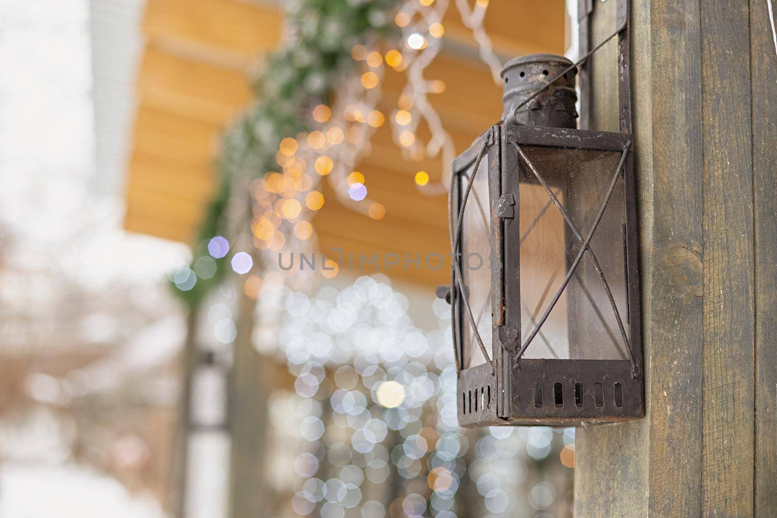 Antique street iron lantern hanging on the wooden post of a house decorated with glowing garlands outside. Close up