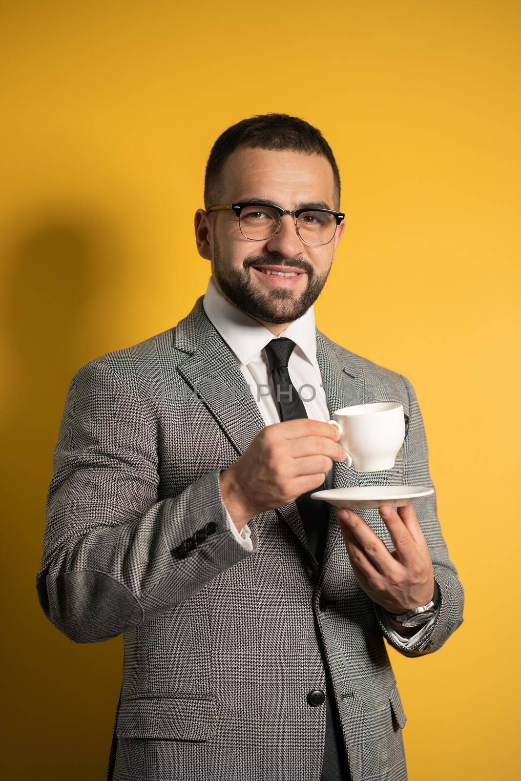 Handsome bearded young man in eye glasses in formal wear holding a cup of coffee isolated on yellow background. Business, hot drinks and people concept.