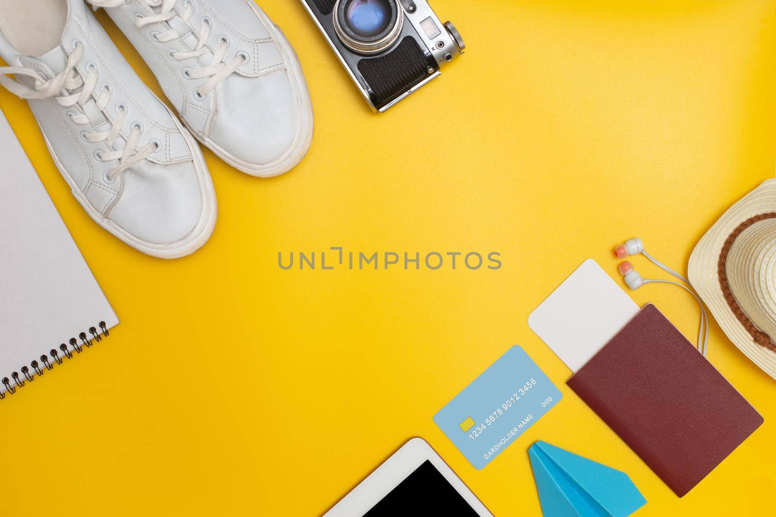 Travel kit: white sneakers, notebook, mobile device, headphones, passport with blank ticket, credit card, paper airplane, straw hat lie in a row diagonally on a yellow background. View from above. Flat lay