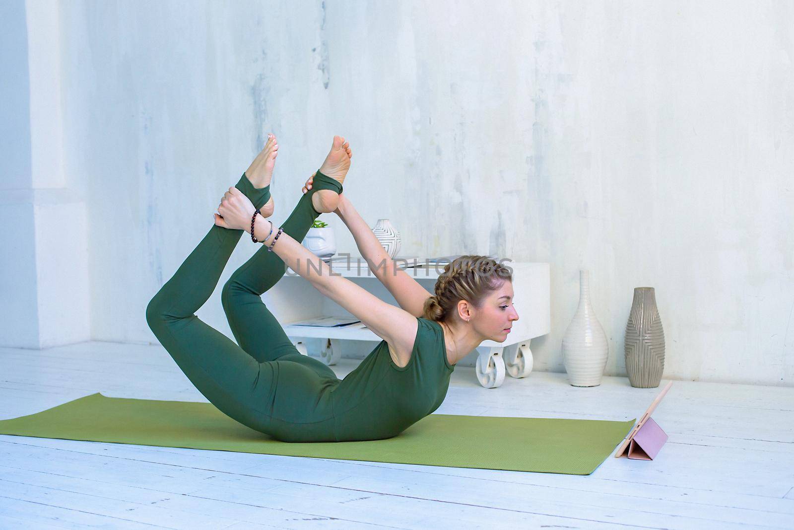 A beautiful and slender girl in green sportswear practices yoga on green mat , in a bow pose, online, using a digital tablet on the floor in a light interior.