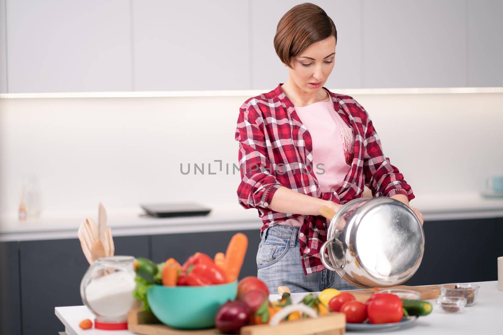Young woman cleans or mixes ingredients in metallic pot with vegetables on the counter. Woman with short hair in a red plaid shirt cooking in a modern kitchen. home lifestyle concept. Vegan concept.