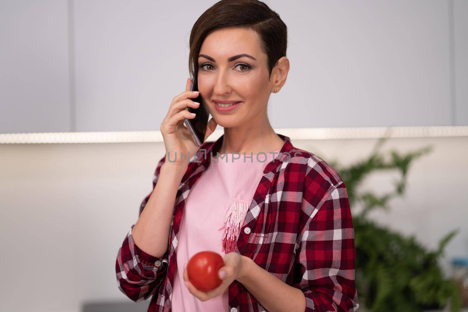 Talking on the phone with tomato in the hand young pretty housewife with a bob hairstyle prepares food in the kitchen. Healthy food at home. Healthy food leaving - vegan concept.