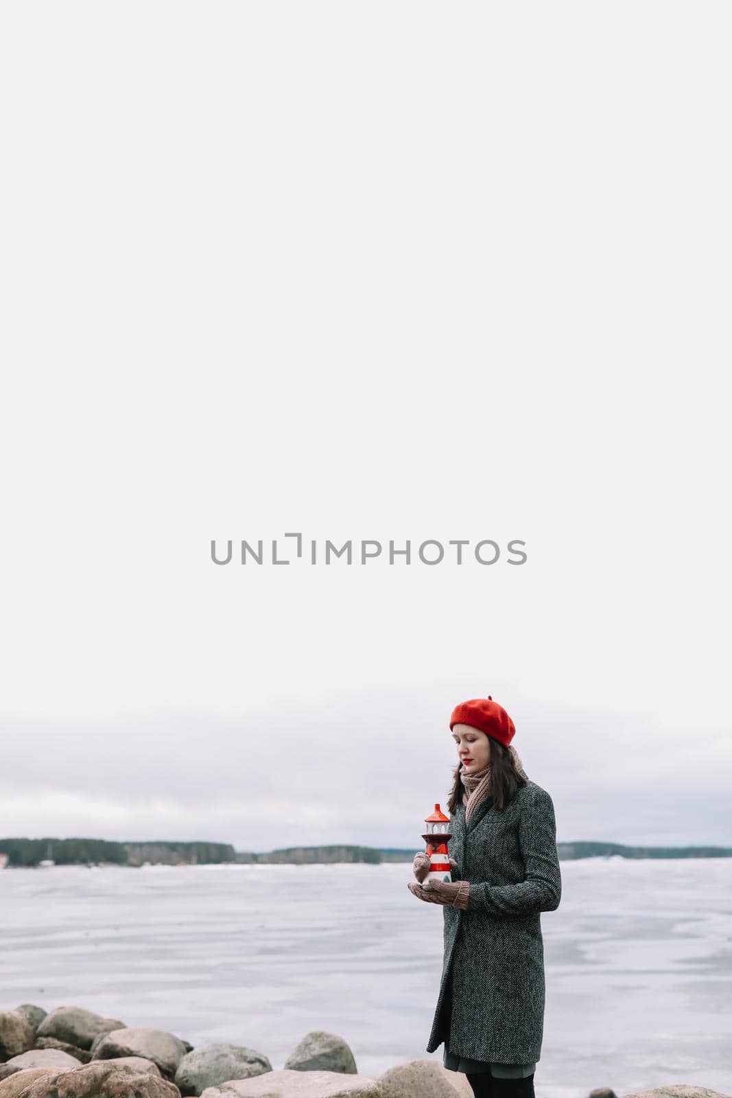 Winter portrait of young woman in a coat and red hat holding decorative lighthouse and standing at the shore of frozen sea. winter, travel, sea background. windy weather, amazing ice seaside