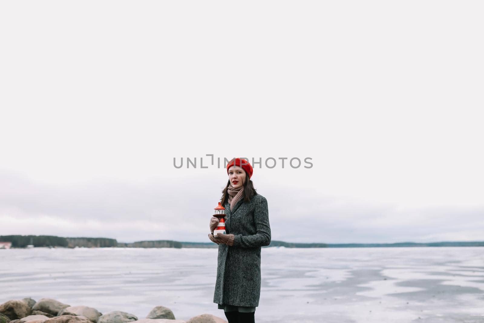 Winter portrait of young woman in a coat and red hat holding decorative lighthouse and standing at the shore of frozen sea. winter, travel, sea background. windy weather, amazing ice seaside