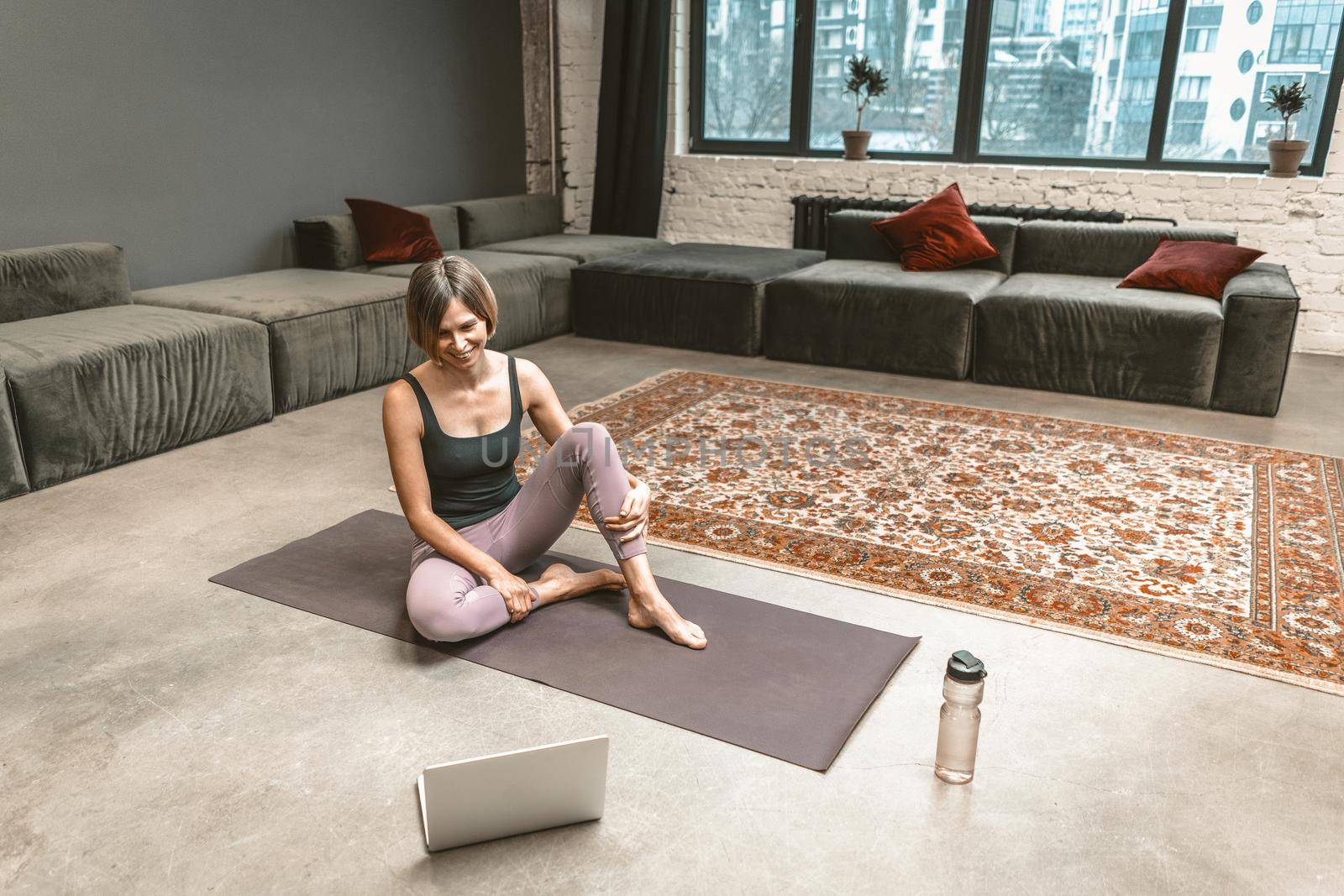Sporty Woman Sits on a Sport Mat and Has an Online Video Chat with her Fitness Coach on Laptop During the Quarantine Period. Home Workouts. City Background. Close-up. High quality photo