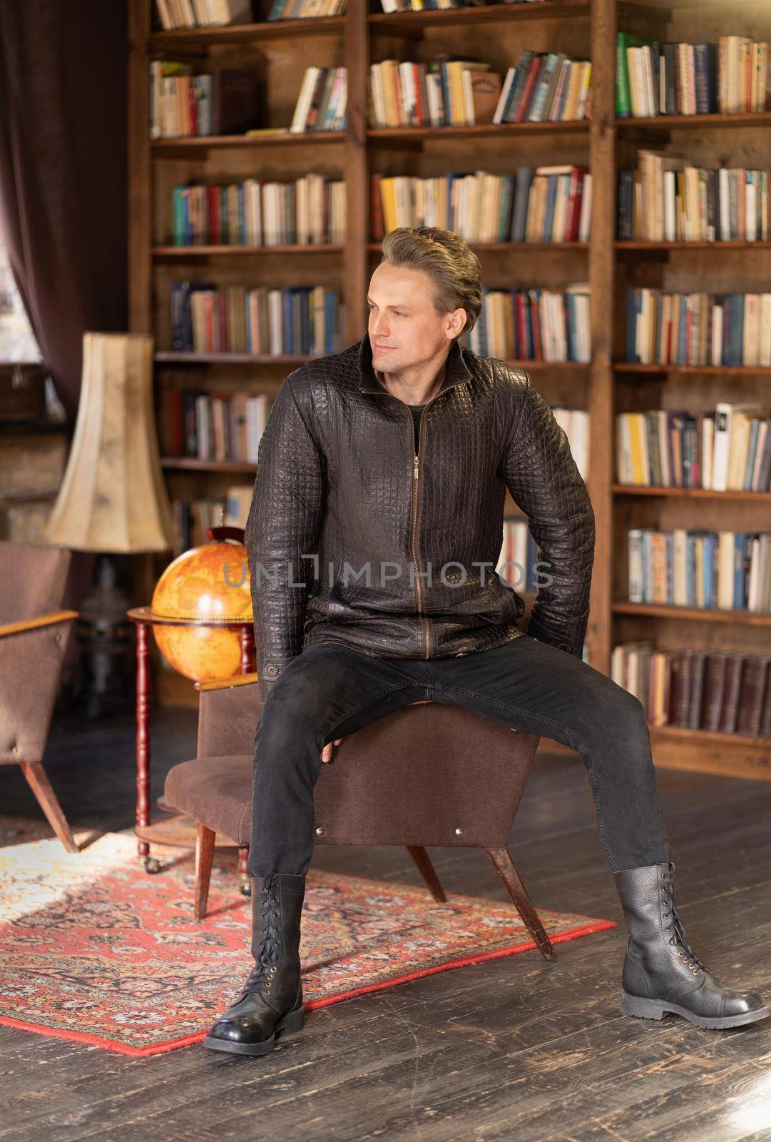 Caucasian Male Model Posing in Loft Studio in Stylish Leather Jacket. Sexy Man Sitting in the Armchair at the full of Books Shelves Background. Close-up Portrait. Book Shelves Background. High quality photo