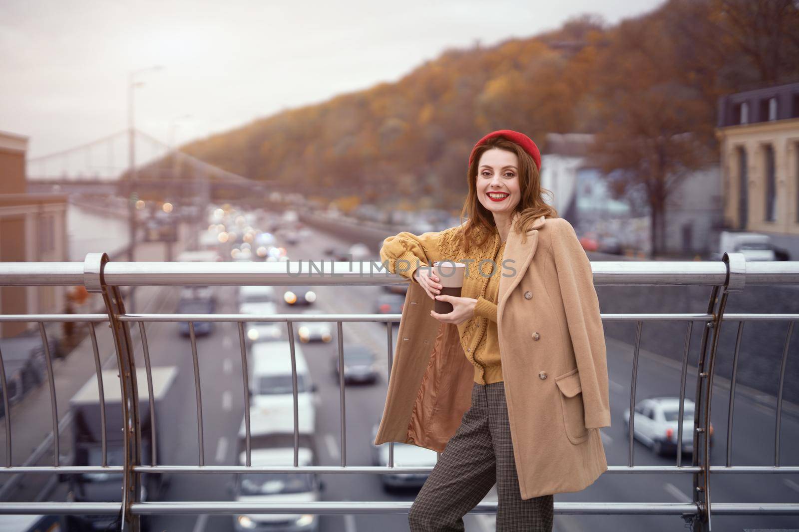 Portrait of a pretty smiling french young woman looking at camera standing at pedestrian bridge with cars on the road. Looking happy fashion girl in a ed beret and beige coat.