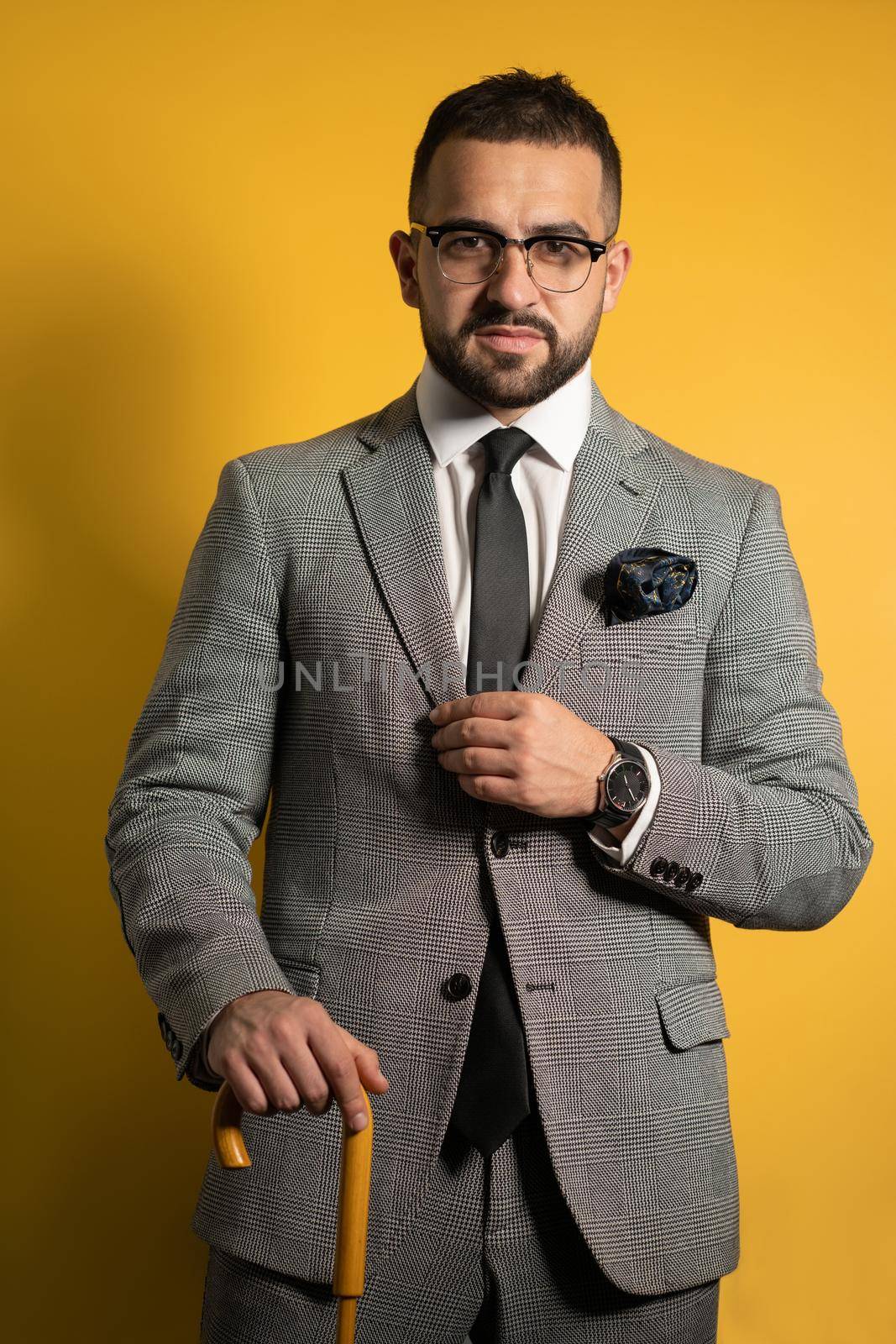 English style handsome young man wearing grey suit with one hand lifted standing with cane or umbrella in his hand isolated on yellow background.