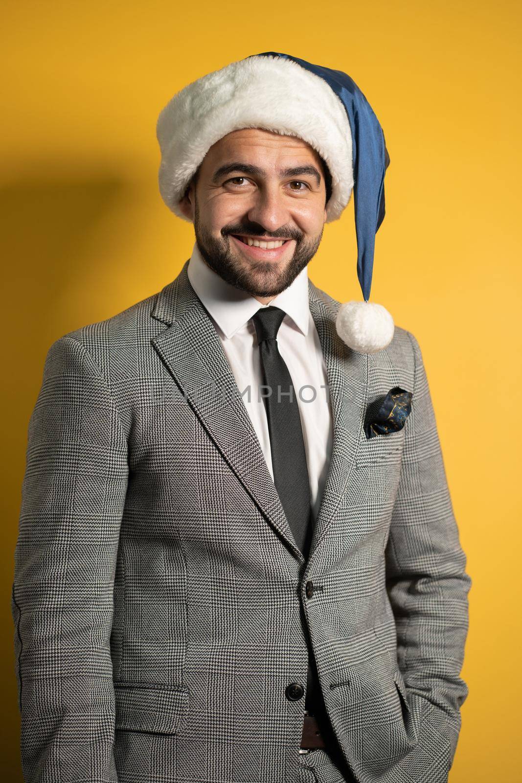 Young handsome bearded smiling Santa man wearing blue Santa Claus hat and suit, looking at camera isolated on yellow background. Positive emotions, facial expressions, feelings seasonal holidays by LipikStockMedia