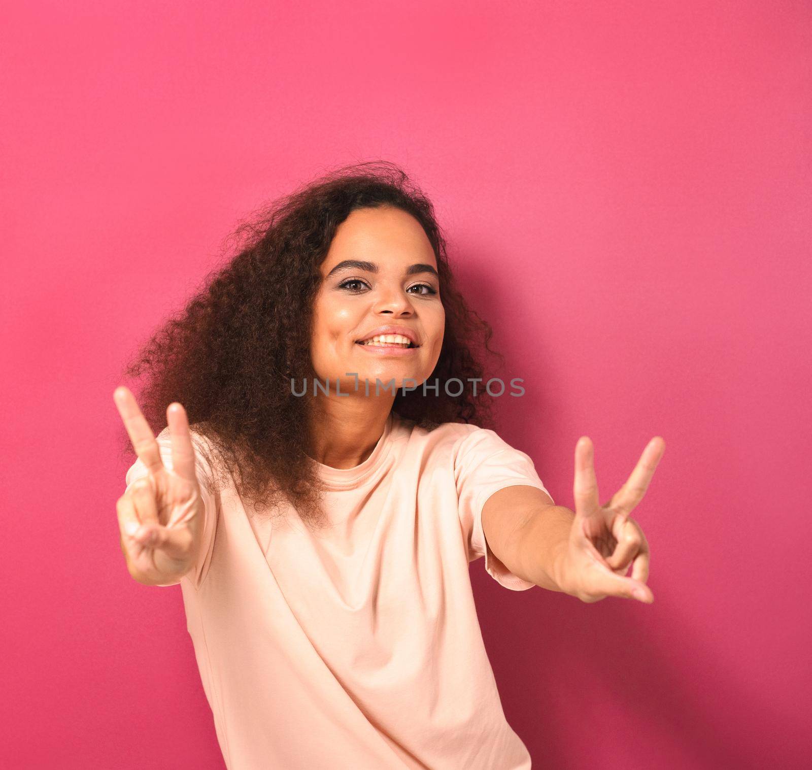 Happy sending PEACE gesture with two hands beautiful young African American woman looking positively at camera wearing peachy t-shirt isolated on pink background. Beauty concept. Square crop.