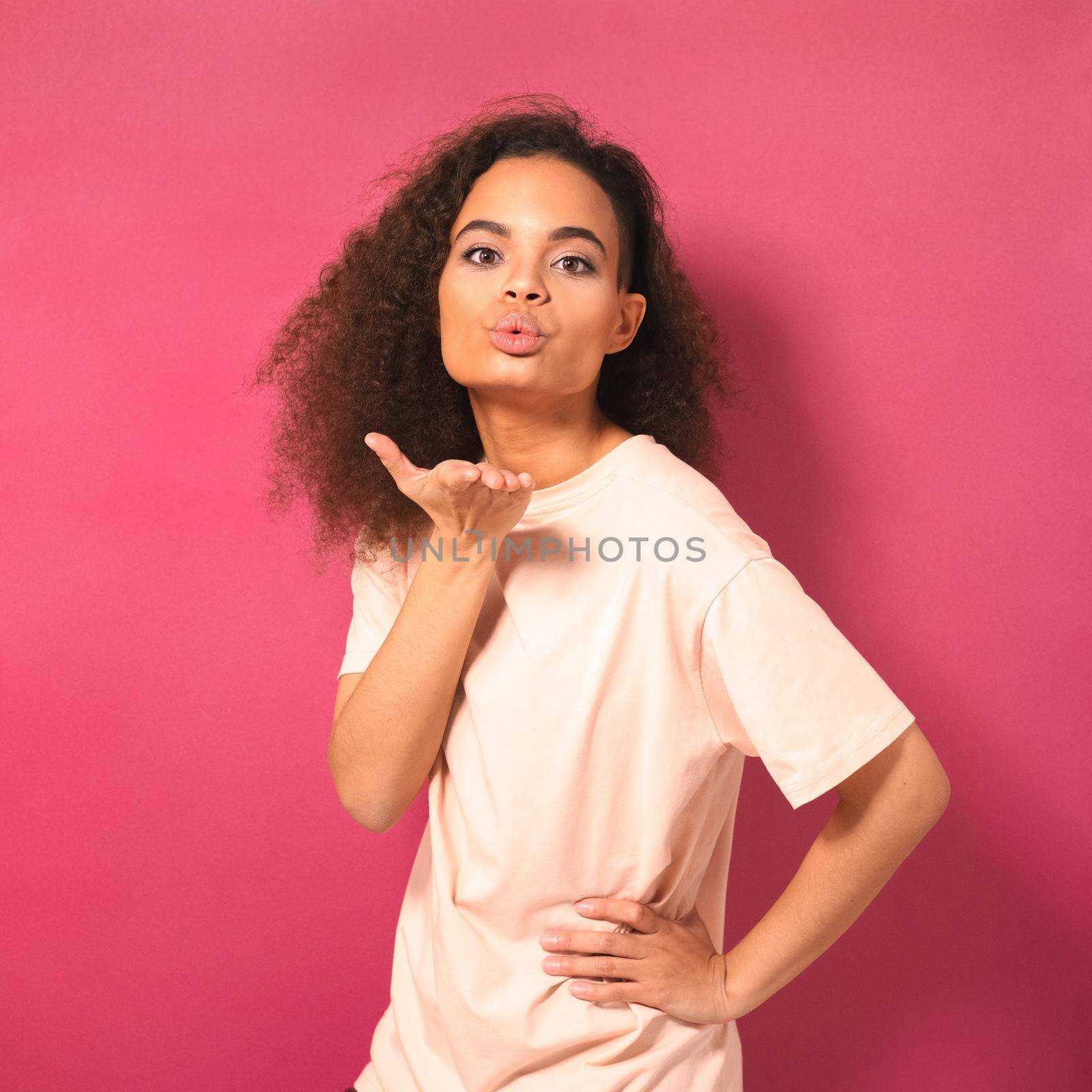 Beautiful young African American woman with Afro hair sending air kisses, looking positively at camera wearing peachy t-shirt isolated on pink background. Beauty concept by LipikStockMedia