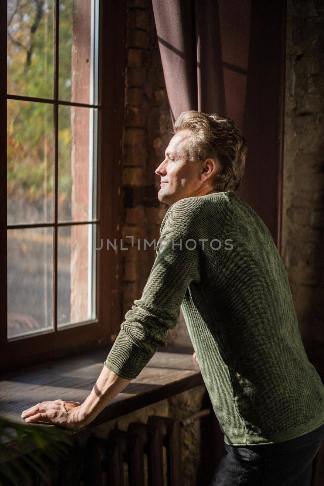 Happy Man Stands at the Window and Looks into the Yard and the Rays of the Sun Shine on his Face. Man Enjoy of Sunny Day. Close-up. High quality photo