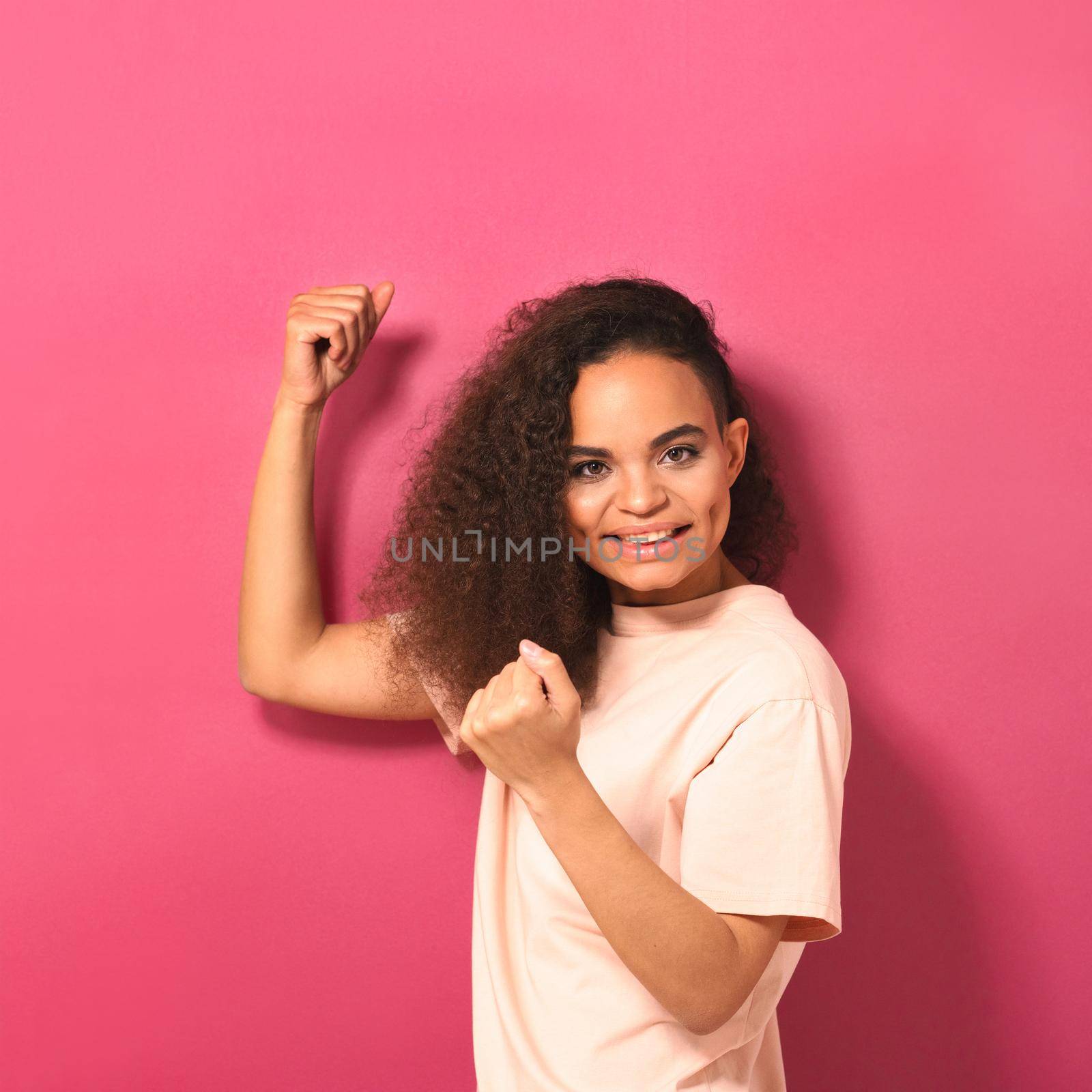 Happy beautiful young African American woman looking positively at camera wearing peachy t-shirt showing her muscles isolated on pink background. Beauty concept. Square crop by LipikStockMedia
