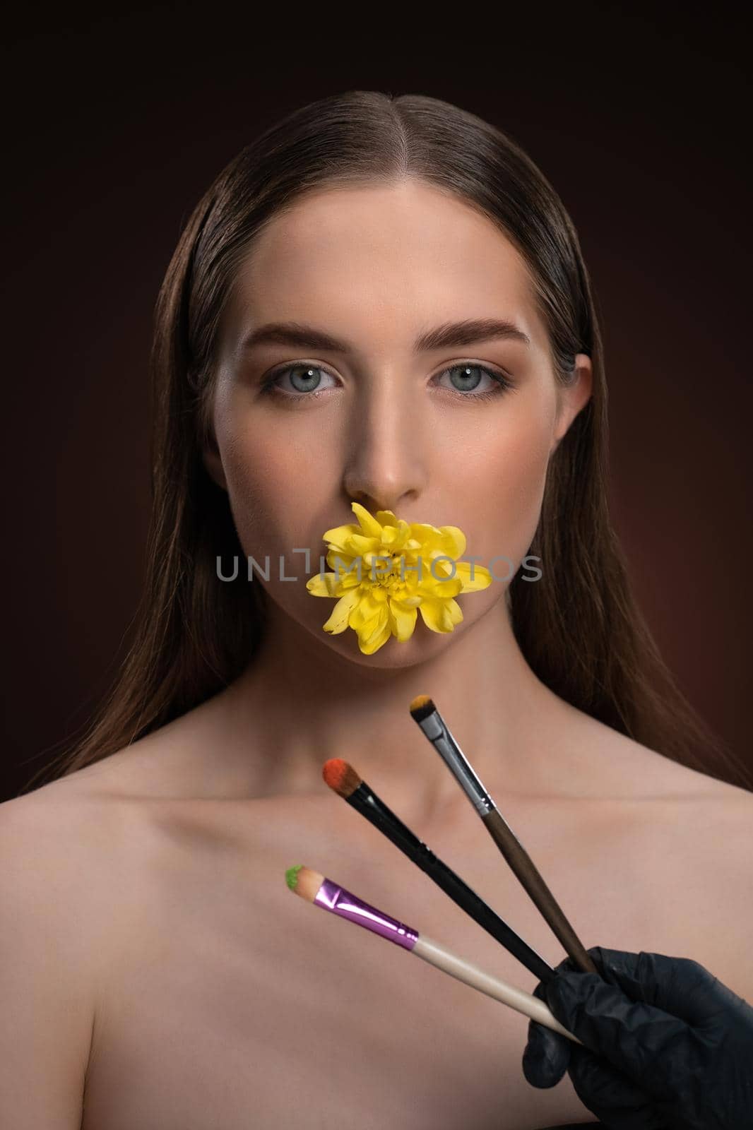 Natural cosmetics concept. Beauty model with bare shoulders holding a flower in her mouth and brushes in front looking at camera isolated on black background. Woman nature cosmetic spa concept by LipikStockMedia