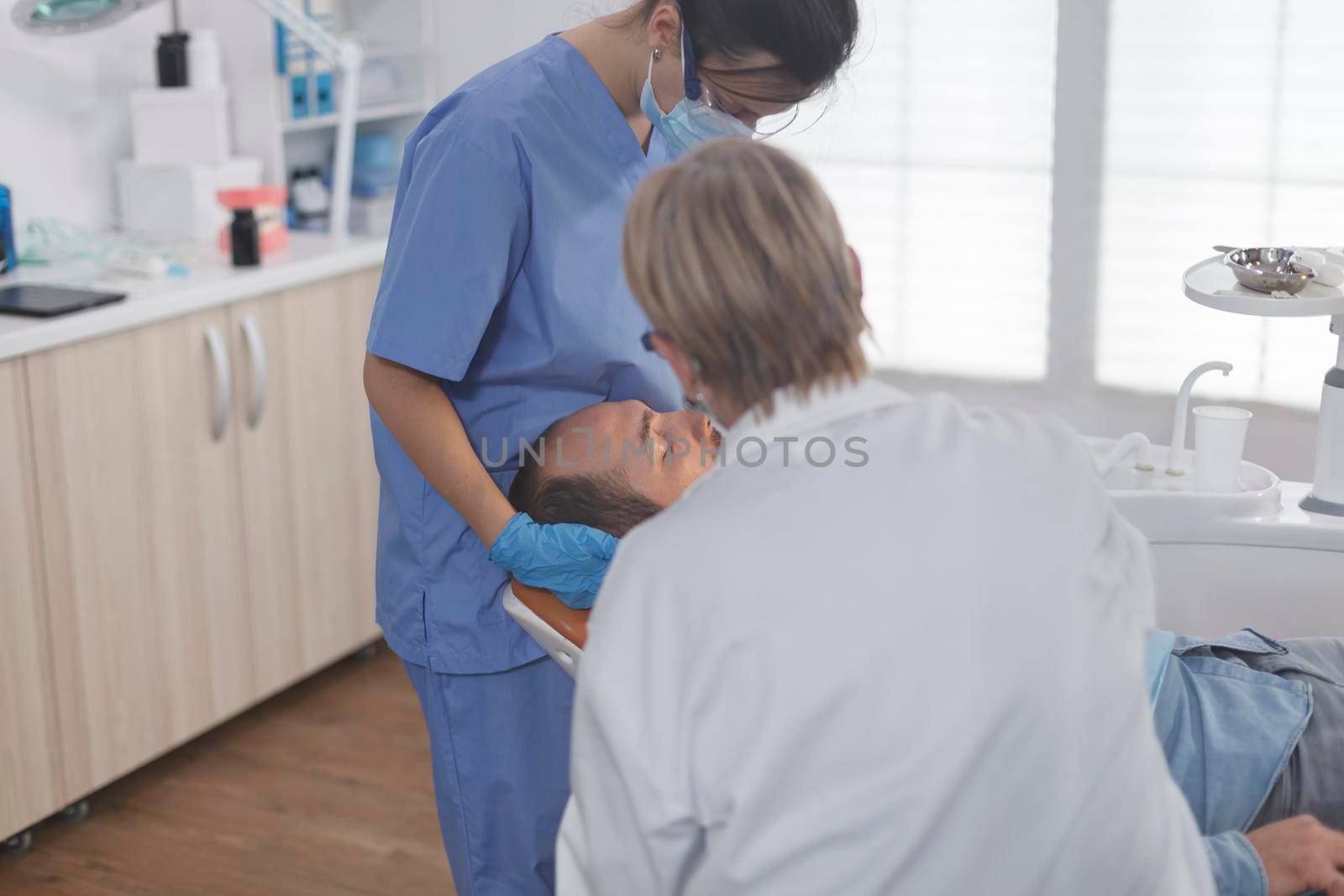 Clinical orthodontist team with face mask examining patient mouth during medical surgery in stomatological office room. Senior woman doctor using professional dentistry tools for cavity procedure