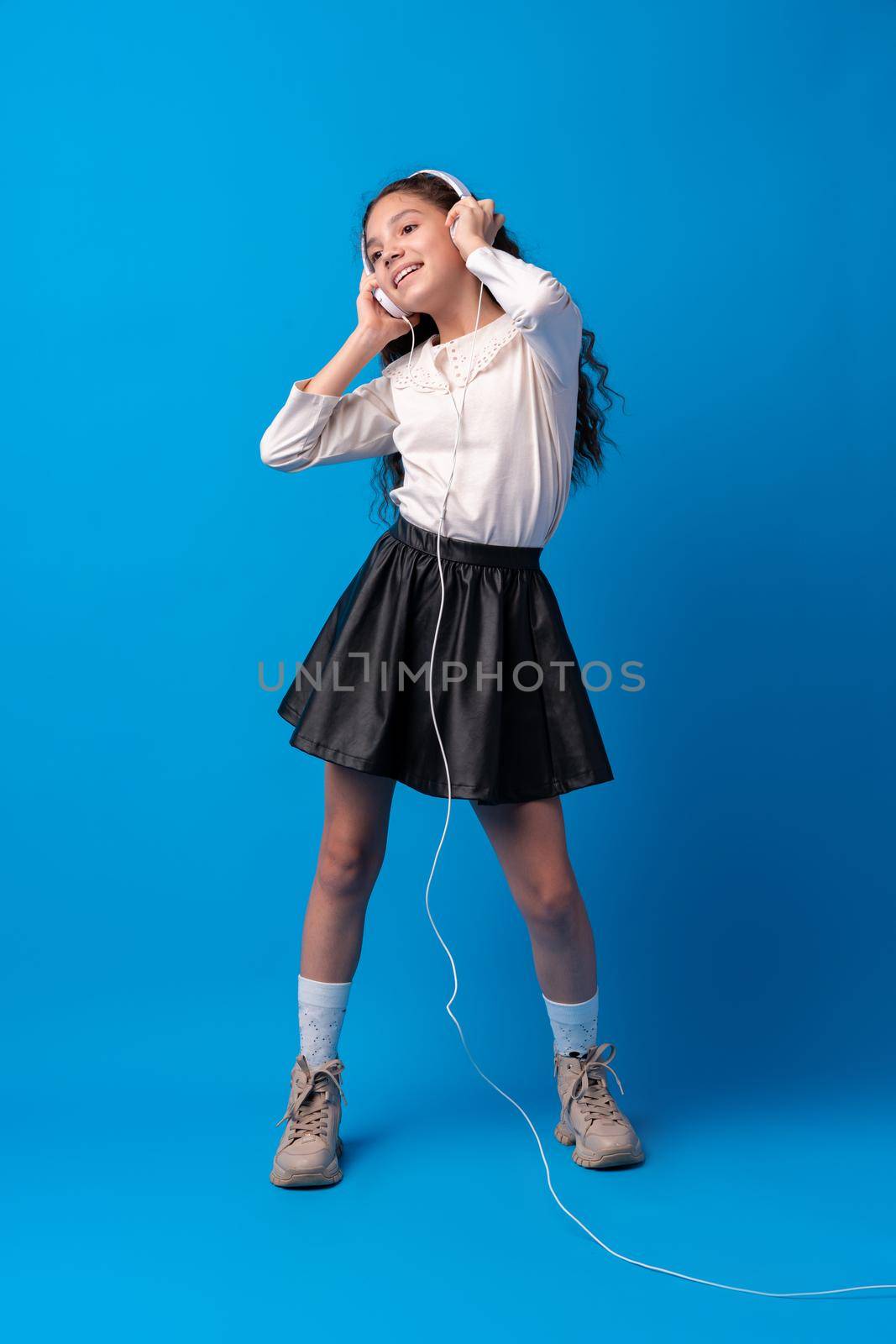 Schoolgirl with headphones listening to music against blue background, portrait