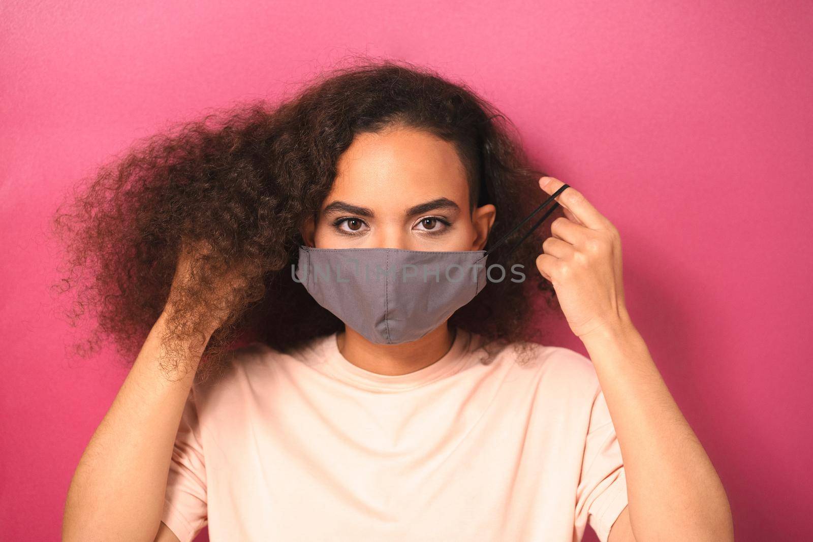 Taking off putting on reusable face mask African American girl in peachy t-shirt, to prevent others from corona COVID-19 and SARS cov 2 infection isolated on pink background.