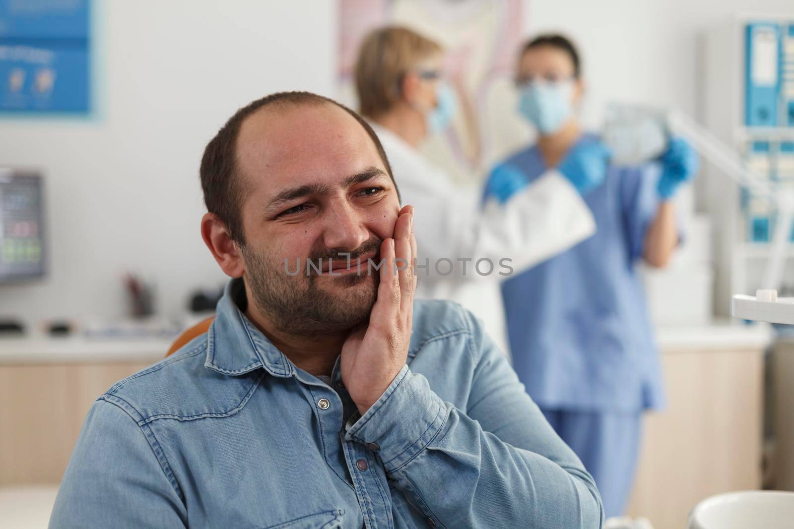 Close up of man patient with toothache sitting on dental chair during stomatological examination waiting for medical expertise. In background specialist team discussing dentistry treatment