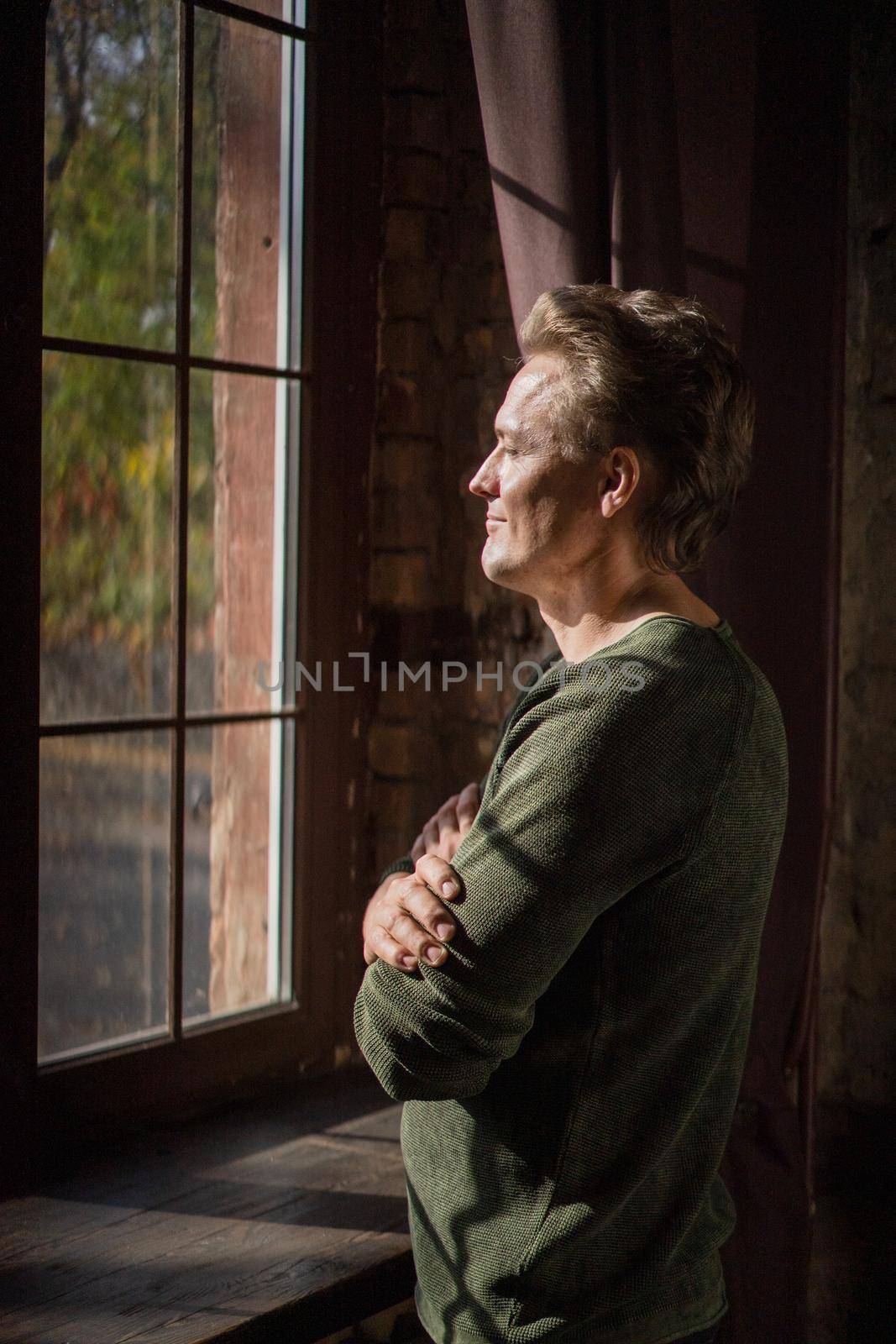 Lonely Man in a Casual Sweater Stands by the Window and Looks at Window in his House. Handsome Man Looks out the Window his Face is IIluminated by the Sun. Close-up by LipikStockMedia