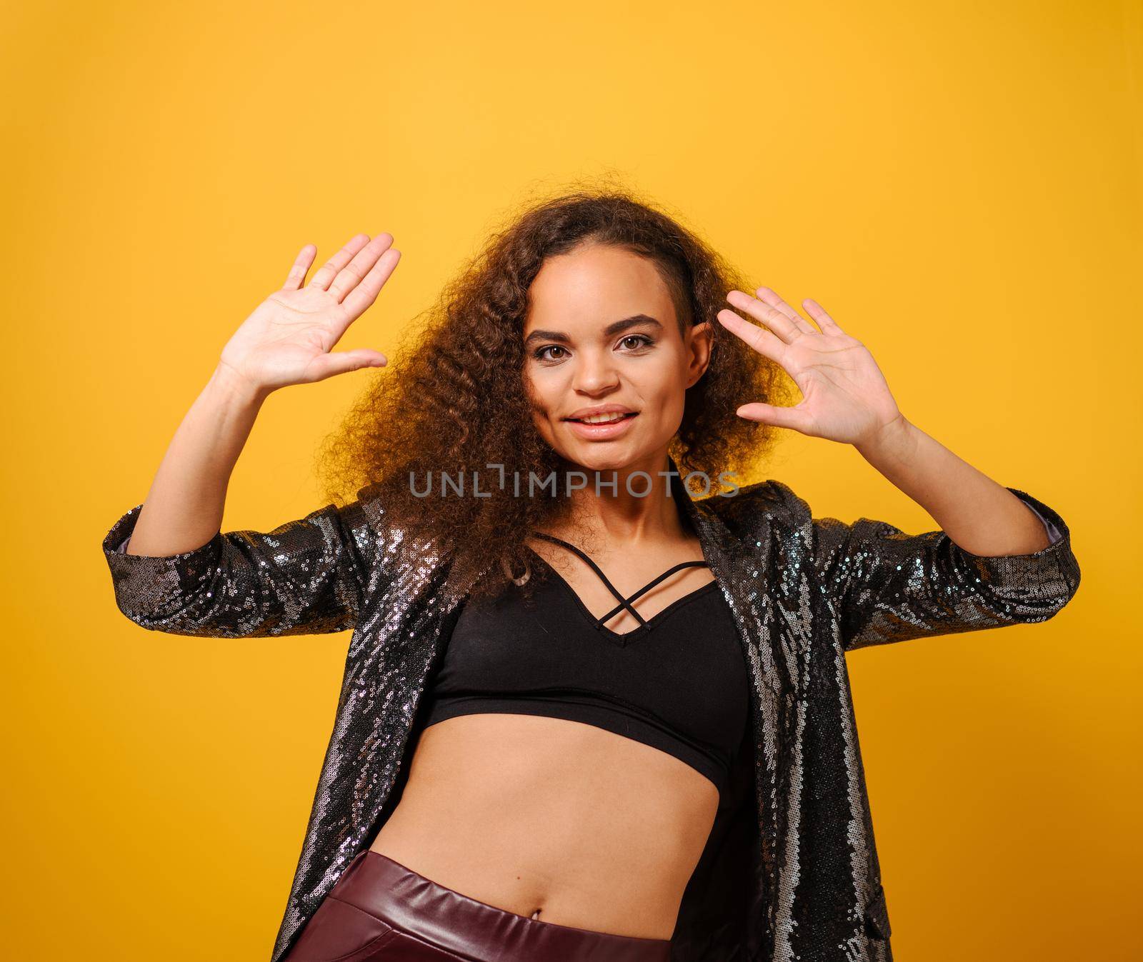 Fashion beautiful African American model young woman with long curly hair posing dancing on camera with hands up wearing shiny jacket and black top. Photo of young african woman on yellow background.