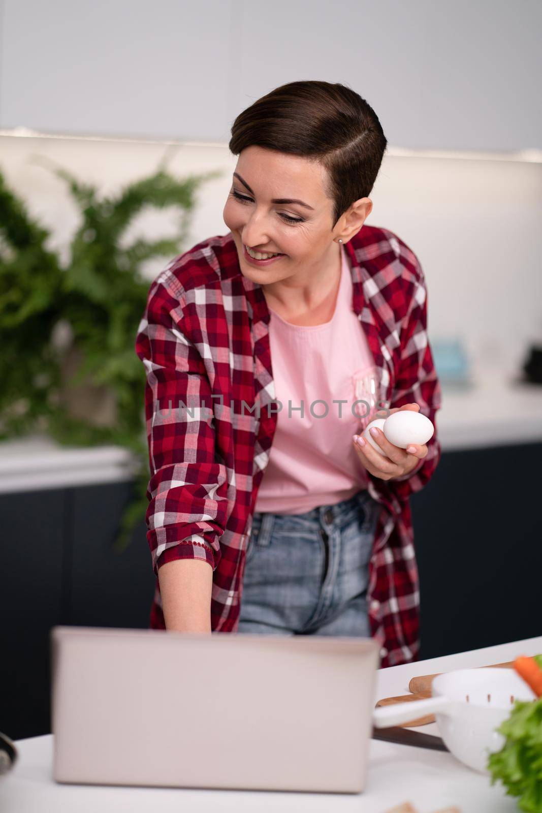Woman in kitchen using laptop holding eggs in hand smiling while reading recipe or having a video call. Woman in red plaid shirt in modern kitchen interior.
