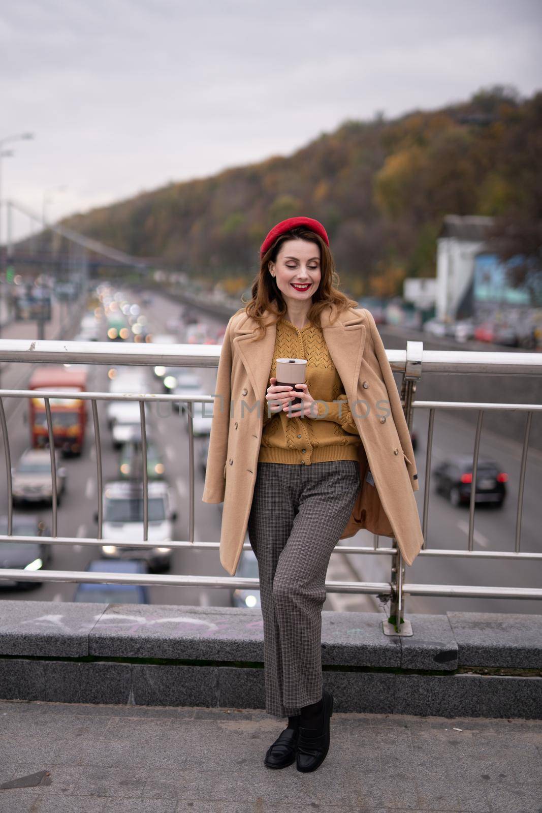 Charming french young woman drinks coffee using a coffee mug looking at camera smiling standing at pedestrian bridge with cars on the road. Looking happy fashion girl in a red beret and beige coat.