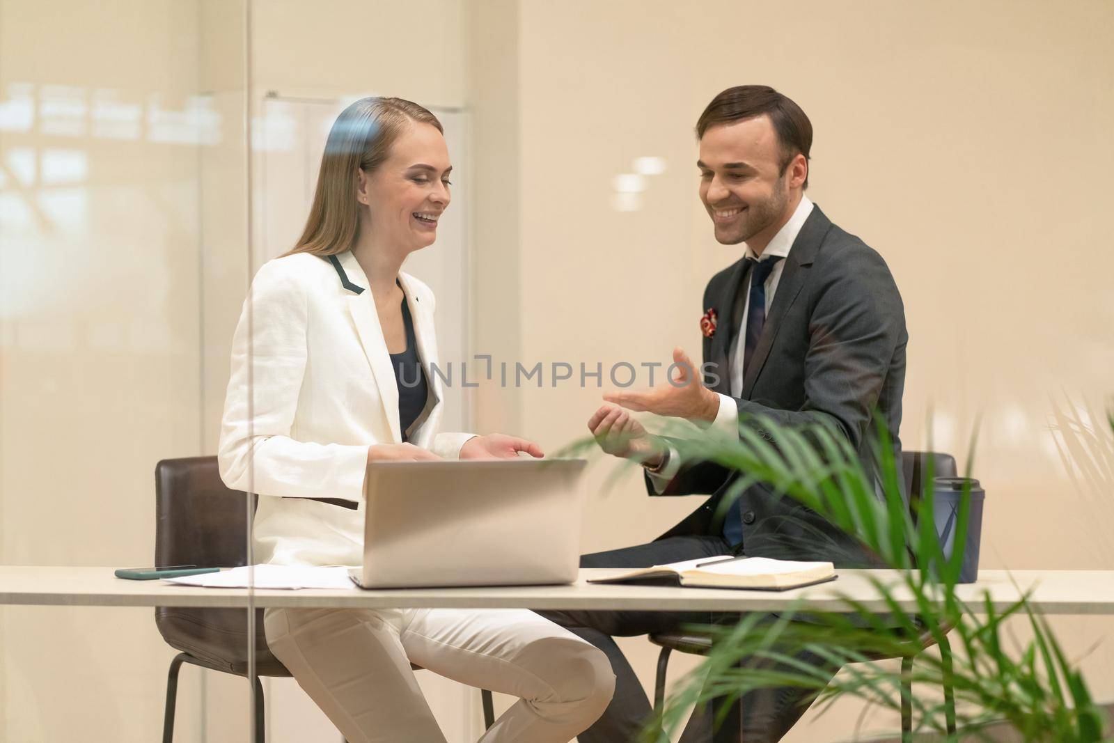 Two laughing project managers discuss updates while having a coffee break. Young business people in the suits with coffee in hands looking down standing behind the glass.
