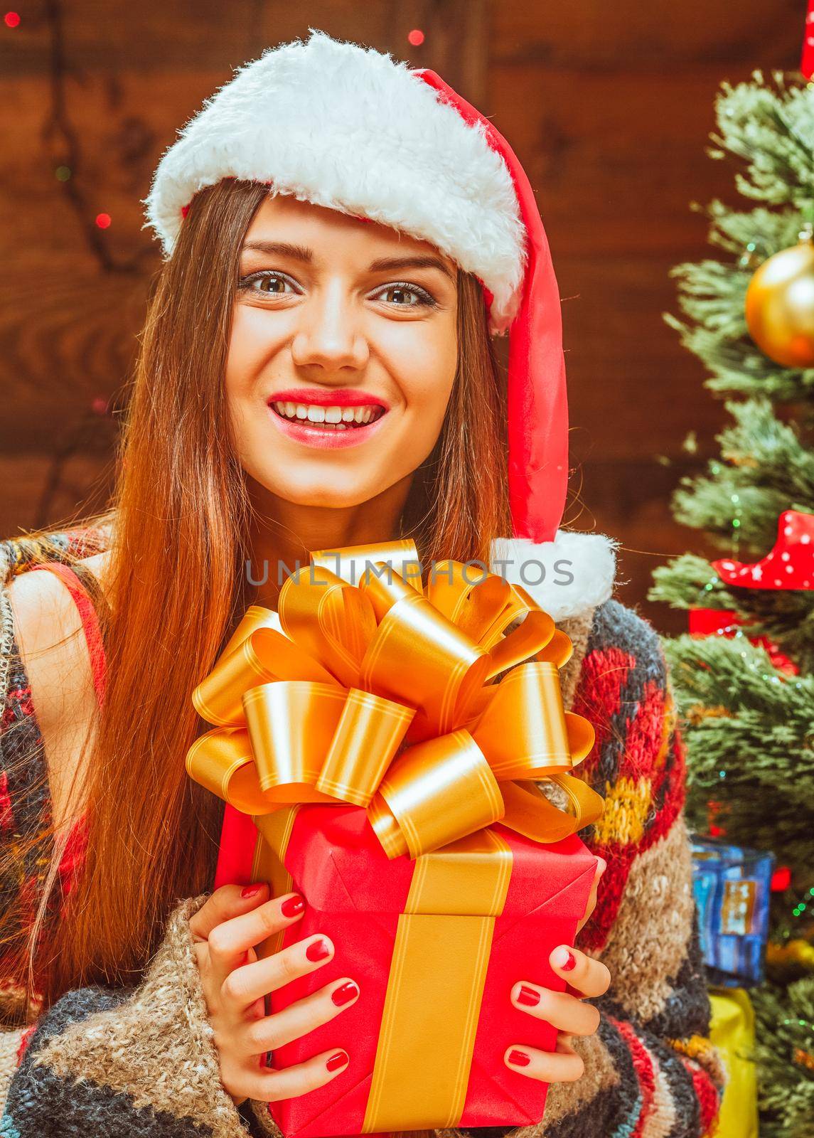 Young Woman in a Knitted Winter Jacket and a Winter Red Hat Holds in her Hands a Red Square Gift Box Tied with a Ribbon and Decorated with a Lush Golden Bow. Christmas and New Year Holidays. Close-up Portrait . High quality photo