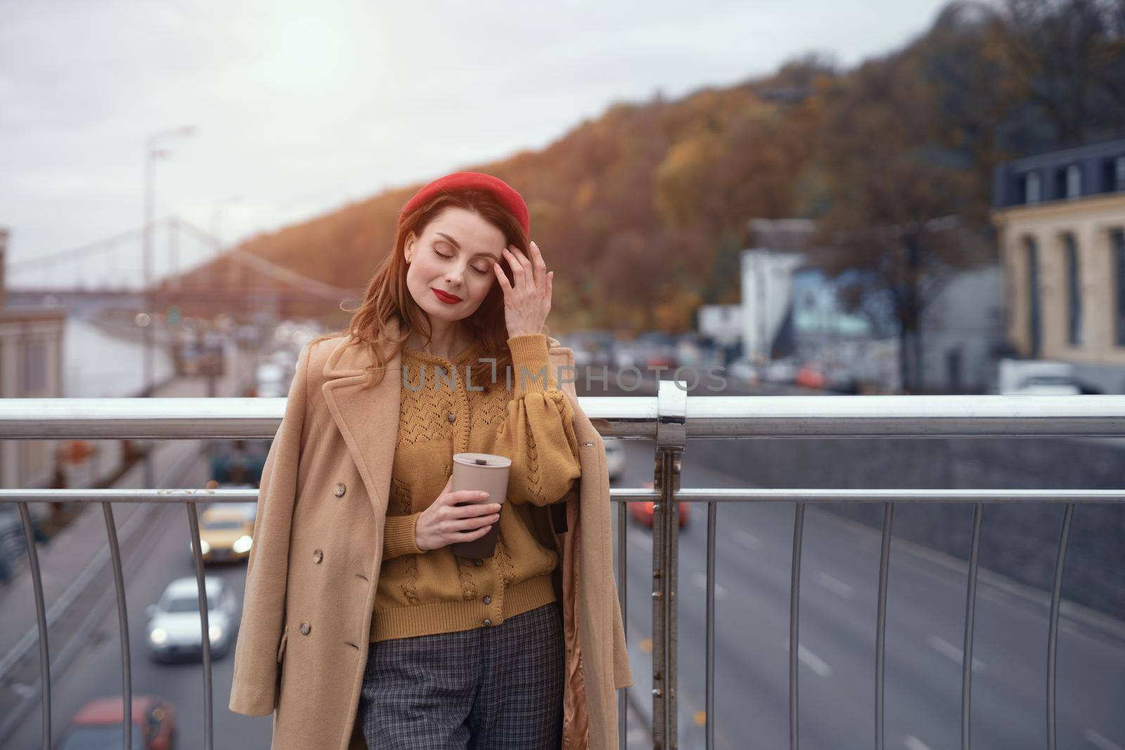 Woman with coffee mug on city street. Charming woman enjoying her cup of coffee standing in a spring urban bridge with traffic behind. French woman in red beret with background of urban city.