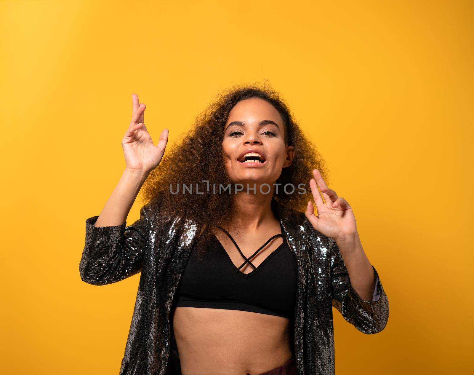 Showing happiness in dancing moves ethnic model girl with beautiful hairstyle smile looking at camera wearing shiny black jacket and black top. Happy young girl on yellow background. Studio shot.